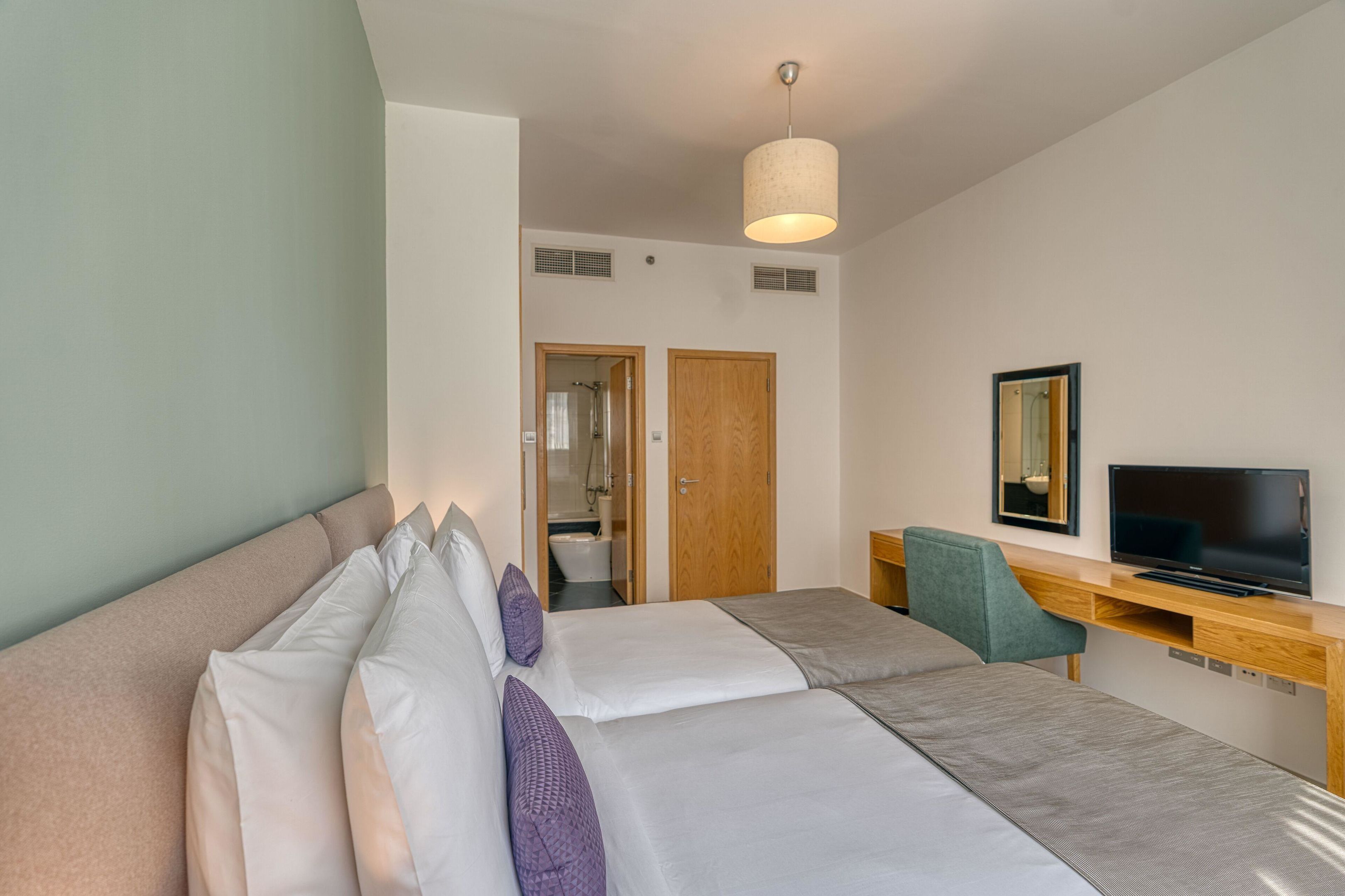 A hotel room with a bed, a desk with a TV, and a bathroom visible through an open door.