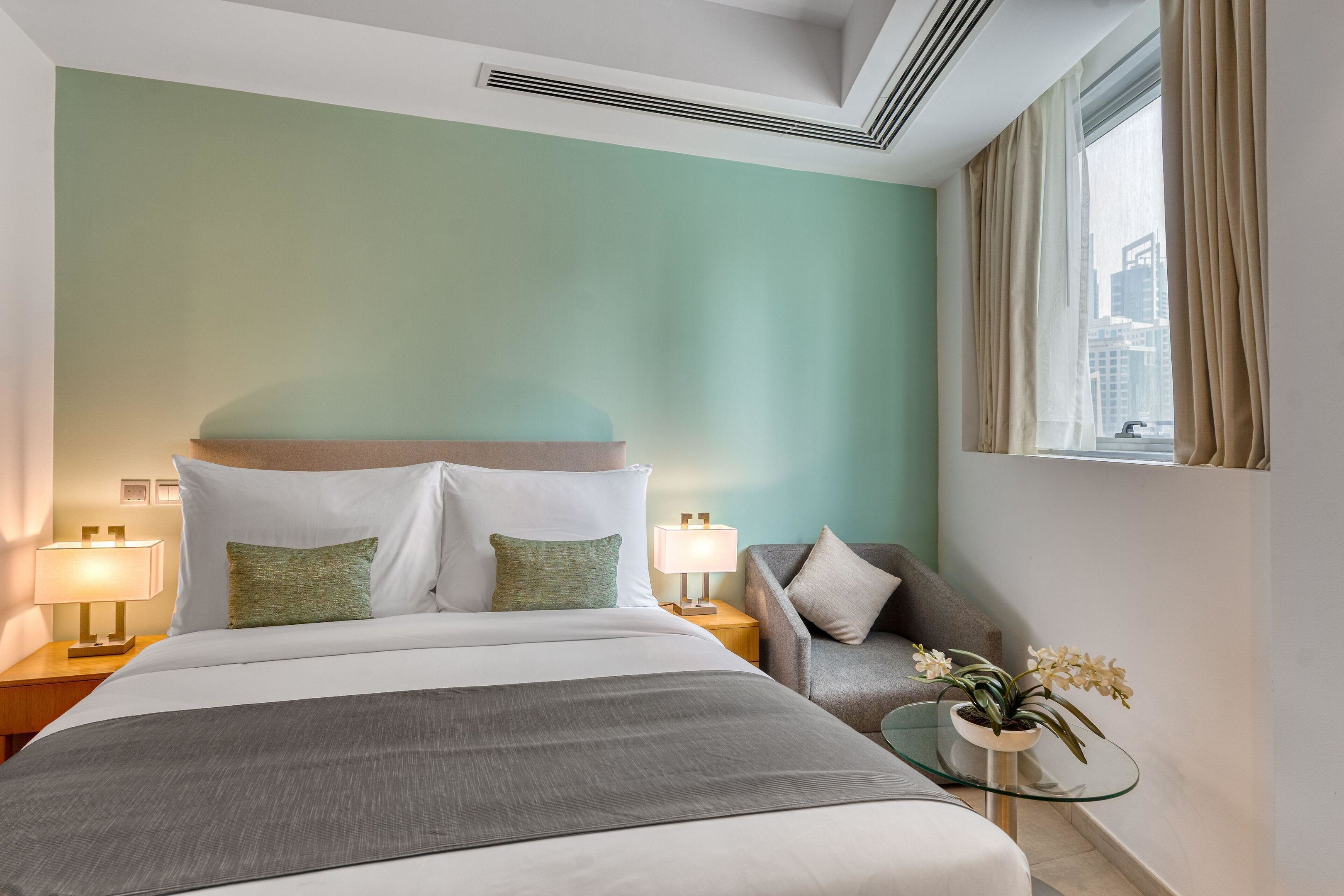 Une chambre d’hôtel avec un lit, une table de chevet avec une lampe, une chaise, une petite table avec une plante et une fenêtre avec des rideaux.