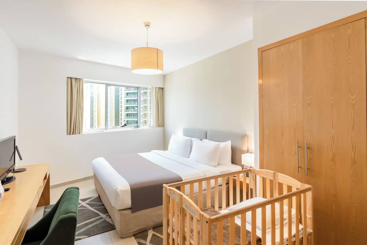 Une chambre d’hôtel comprenant un lit, un berceau, un bureau avec un ordinateur, une fenêtre avec des rideaux et une armoire.