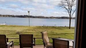 A deck with four chairs and a table, overlooking a lake with a grassy area and a tree.