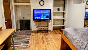 Una habitación con una cama, un escritorio, un televisor sobre un mueble bajo, un reloj en la pared y una planta pequeña.