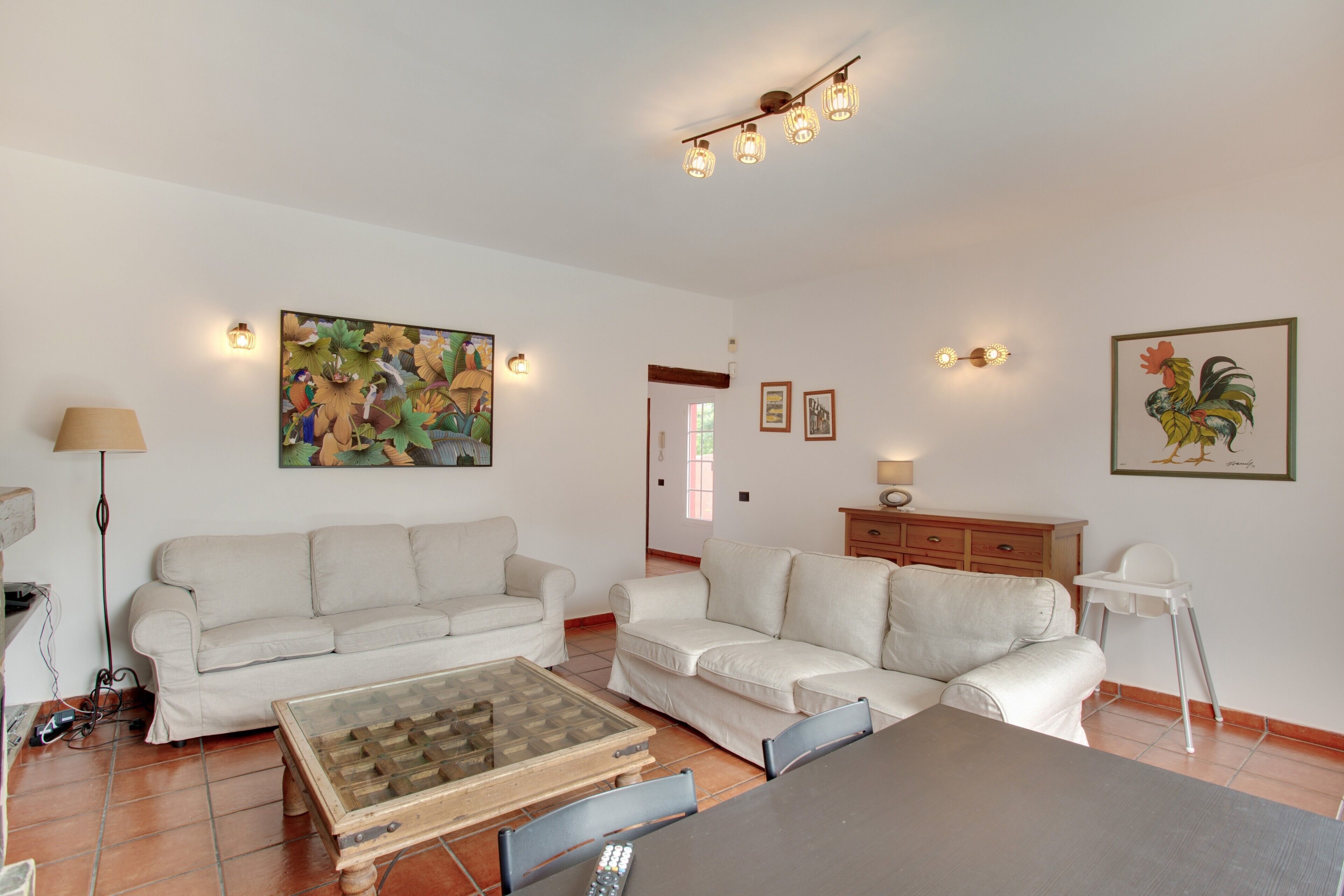 A living room with two white sofas, a coffee table, a dining table, and a painting of a rooster.