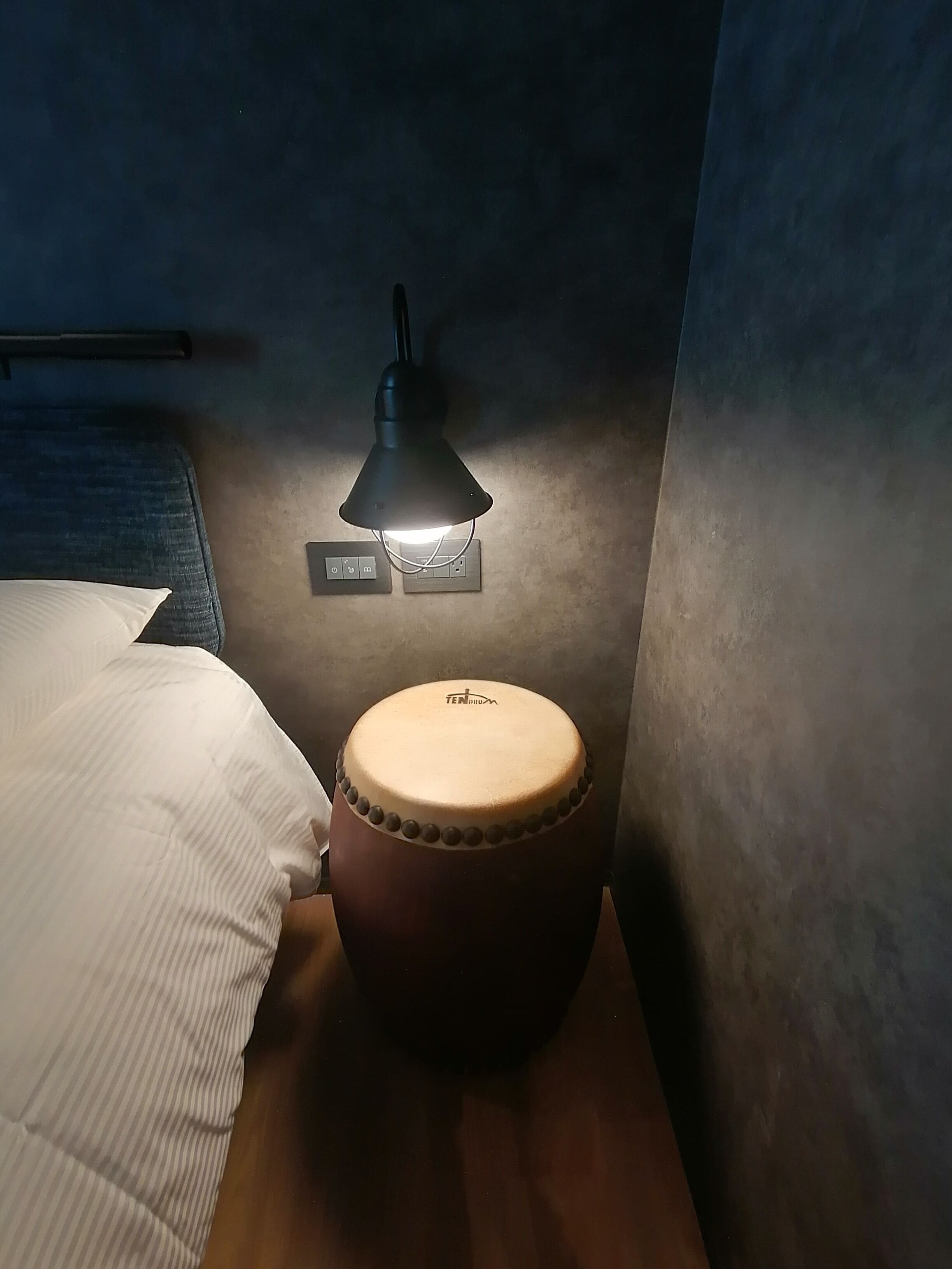 Eine Trommel mit Etikett, eine Steckdose und eine Lampe auf einem Nachttisch in einem Zimmer.