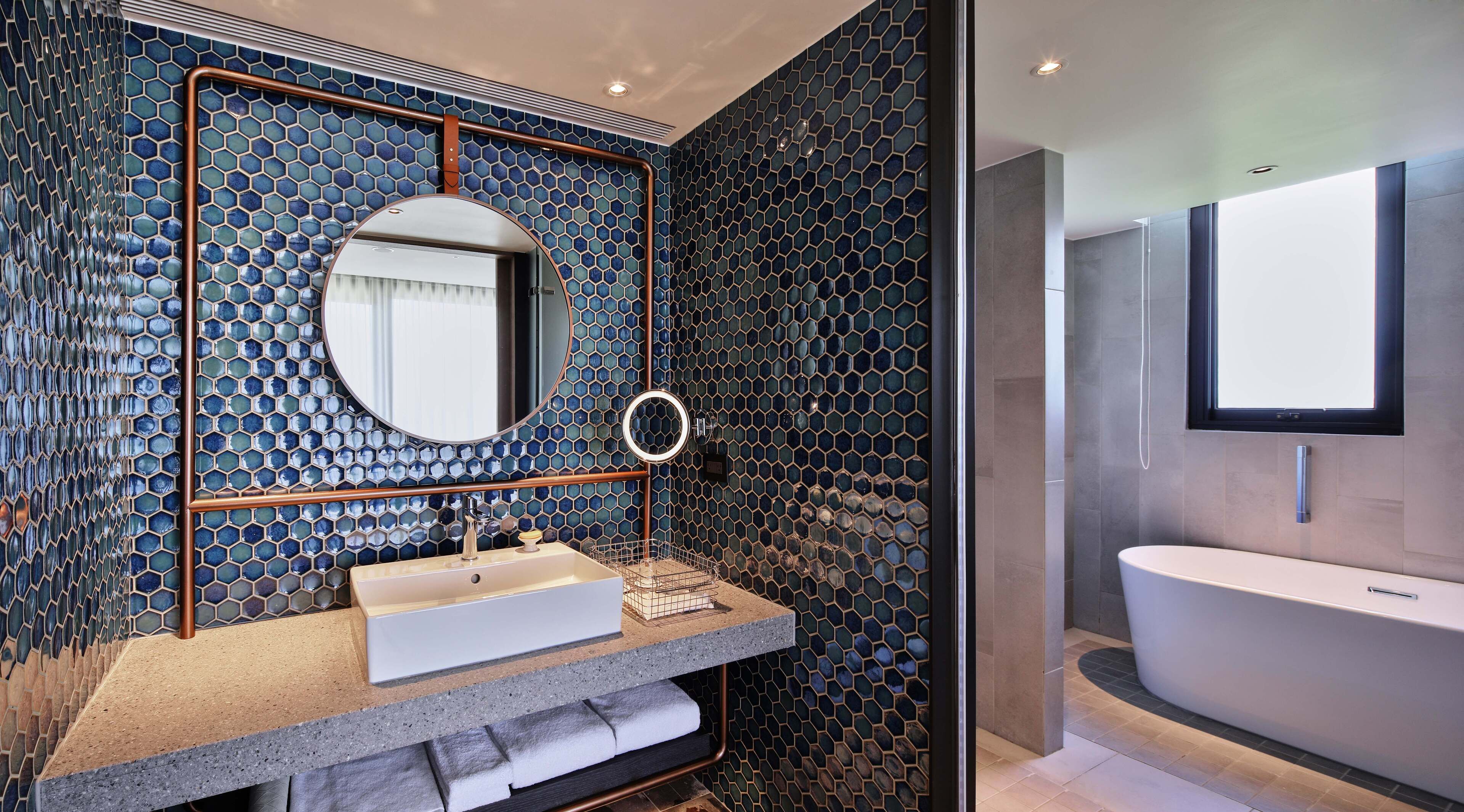 A modern bathroom with a hexagonal mosaic tile wall, a round mirror, a sink with a towel rack, and a standalone bathtub.