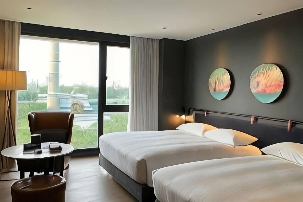A hotel room with two beds, a round table, a chair, and a large window with a view of greenery and a building.