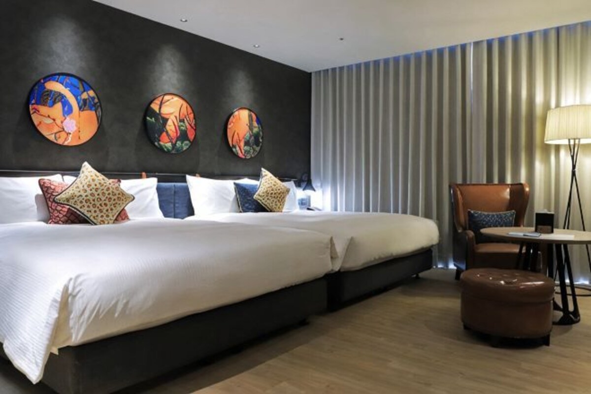A hotel room with a large bed, a chair, a table, and artwork on the wall.