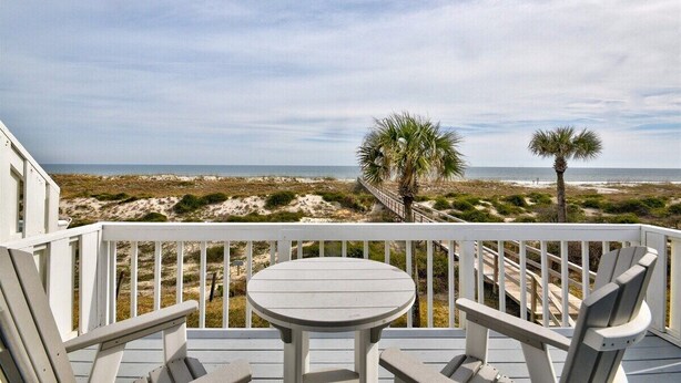 A deck with two Adirondack chairs and a small round table, overlooking a beach with dunes and palm trees. - Anchors Away - Hosted By Moore AI Rentals (Fernandina Beach)