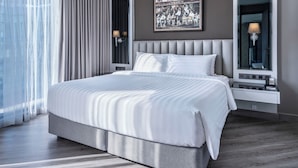 A modern hotel room with a large bed, a bedside table with a lamp, a wall-mounted artwork, and a window with curtains.