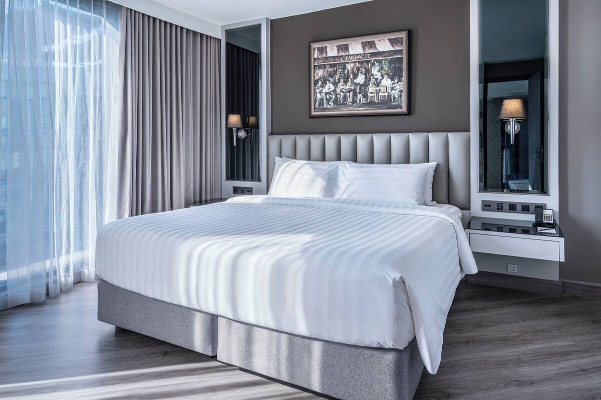A modern hotel room with a large bed, a bedside table with a lamp, a wall-mounted artwork, and a window with curtains.