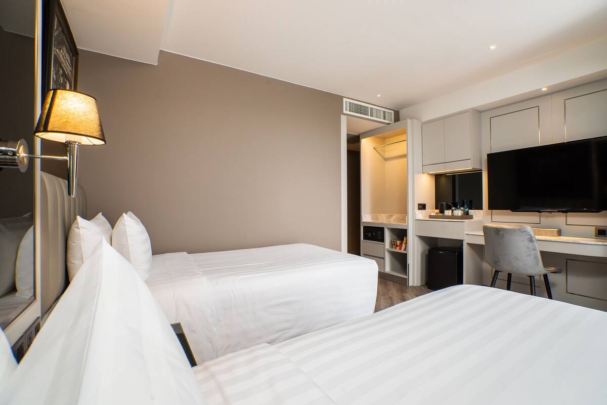 A hotel room with two beds, a desk, a chair, a TV, and a lamp.