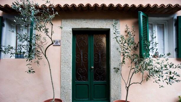 Un edificio con puerta verde, olivos en macetas y un muro de piedra.