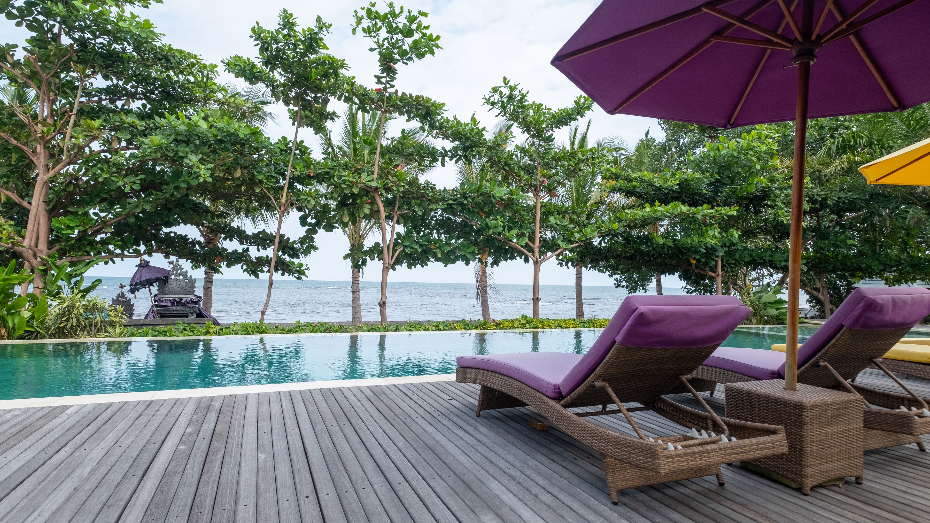 Una terraza de madera con sillones de descanso y una piscina, con vistas a un paisaje tropical con árboles y el océano.
