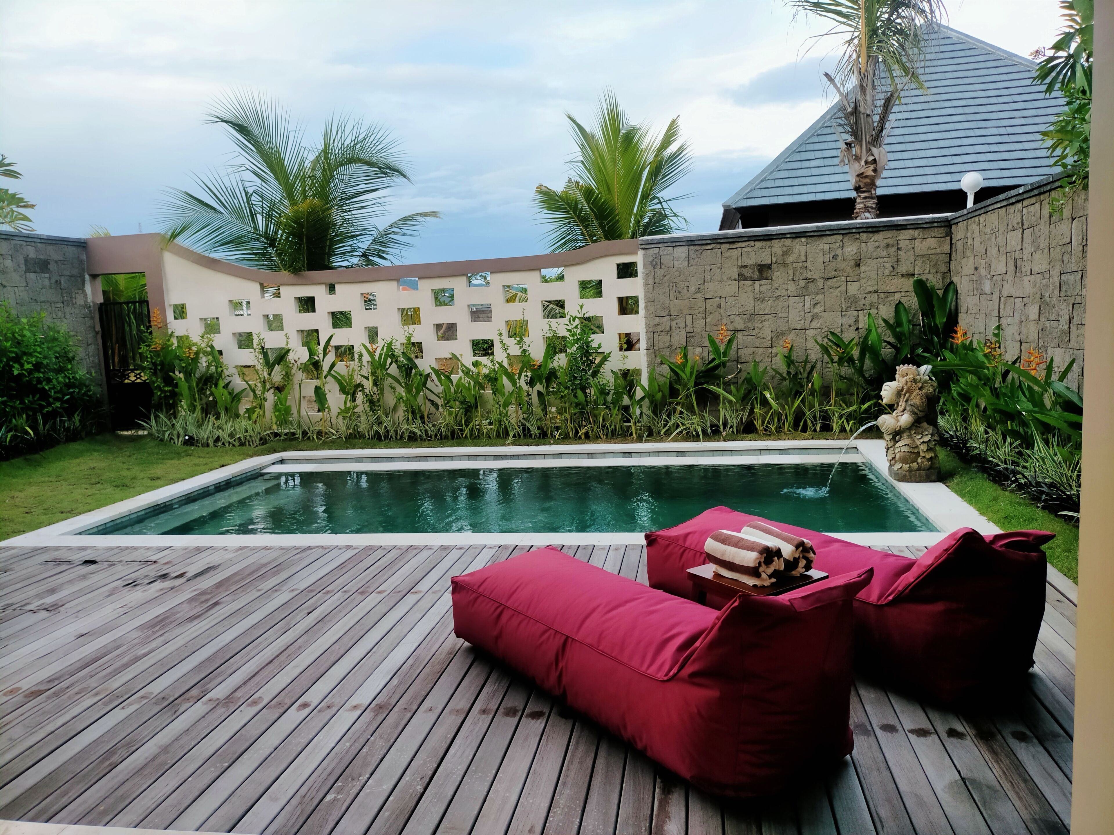 A swimming pool with a wooden deck, surrounded by lush greenery and a stone wall.