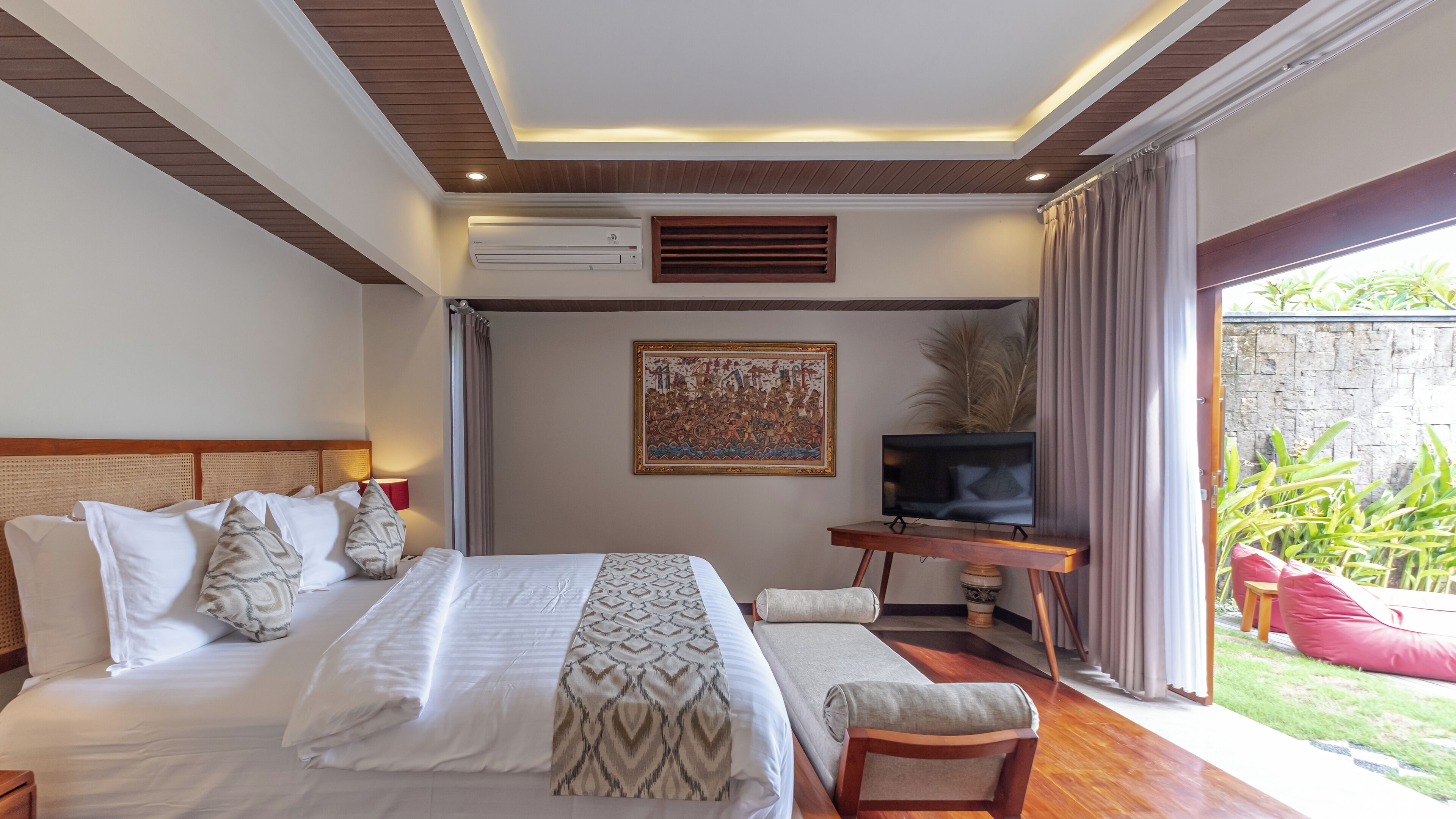 Quarto de hotel com uma cama grande, uma televisão em um suporte de madeira, um quadro na parede e vista para o jardim pela janela.