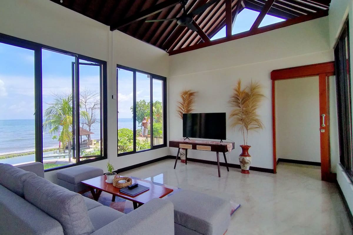 Un salón amplio con un sofá grande, una mesa de centro y un televisor de pantalla plana. Grandes ventanales ofrecen vistas al océano y palmeras.
