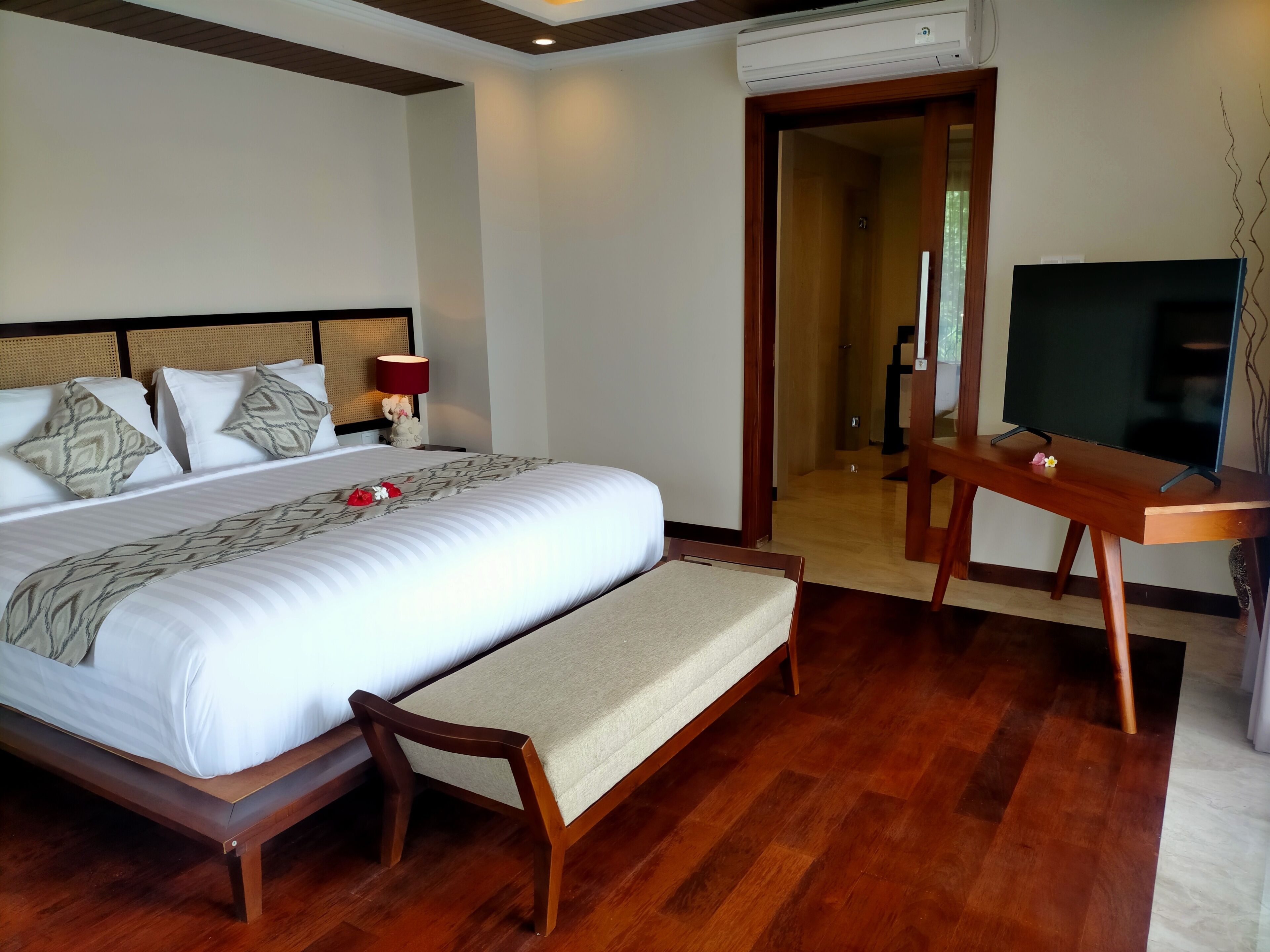 A hotel room with a large bed, a wooden bench, a TV on a wooden stand, and an air conditioning unit on the wall.