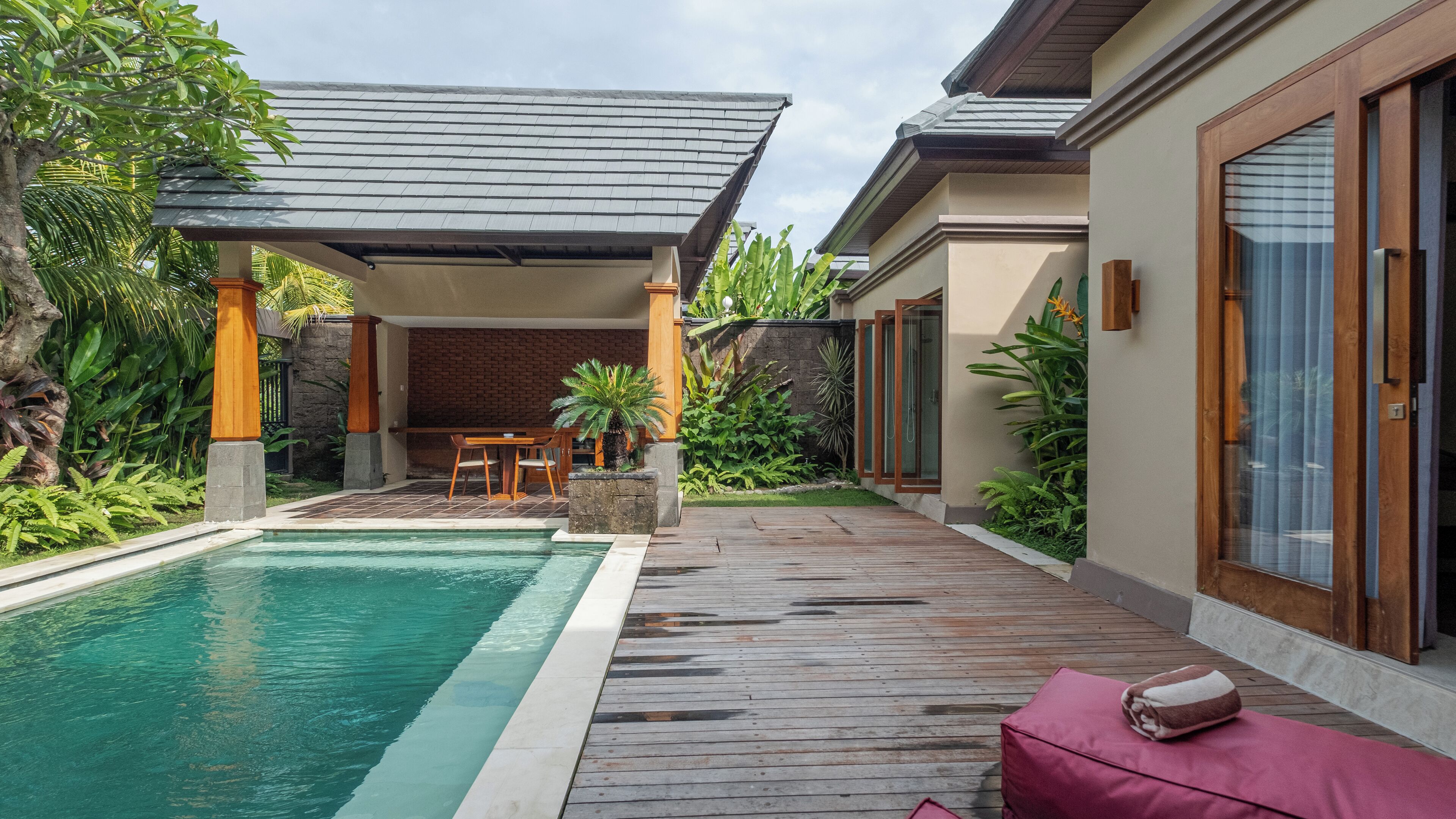 A swimming pool with a wooden deck, a covered seating area, and lush greenery.