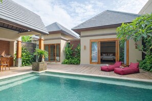 A poolside area with a wooden deck, a seating area with cushions, and a view of a bedroom through a glass door. - The Grand Villandra Resort (Buleleng)
