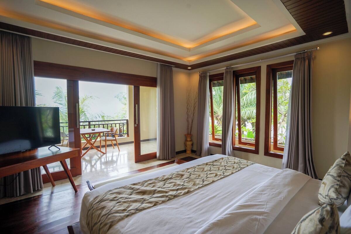 Habitación de hotel con una cama grande, televisor, balcón con mesa y sillas, y ventanas amplias con cortinas.