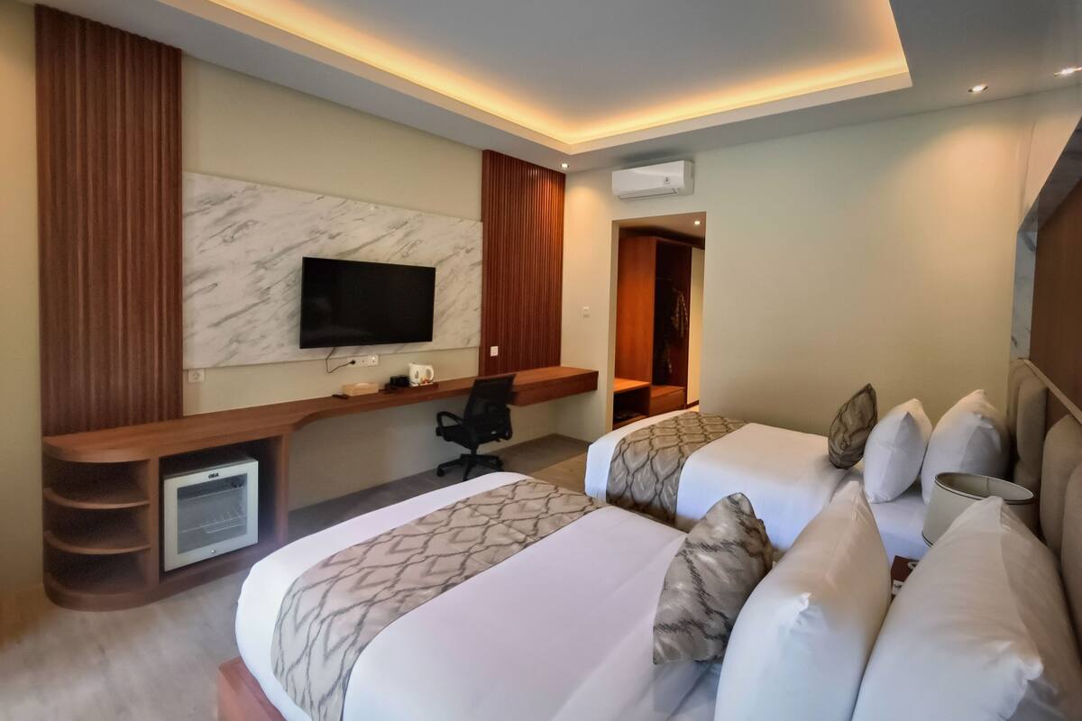 Habitación de hotel con dos camas, un televisor montado en la pared, un escritorio con una silla y una pequeña kitchenette.