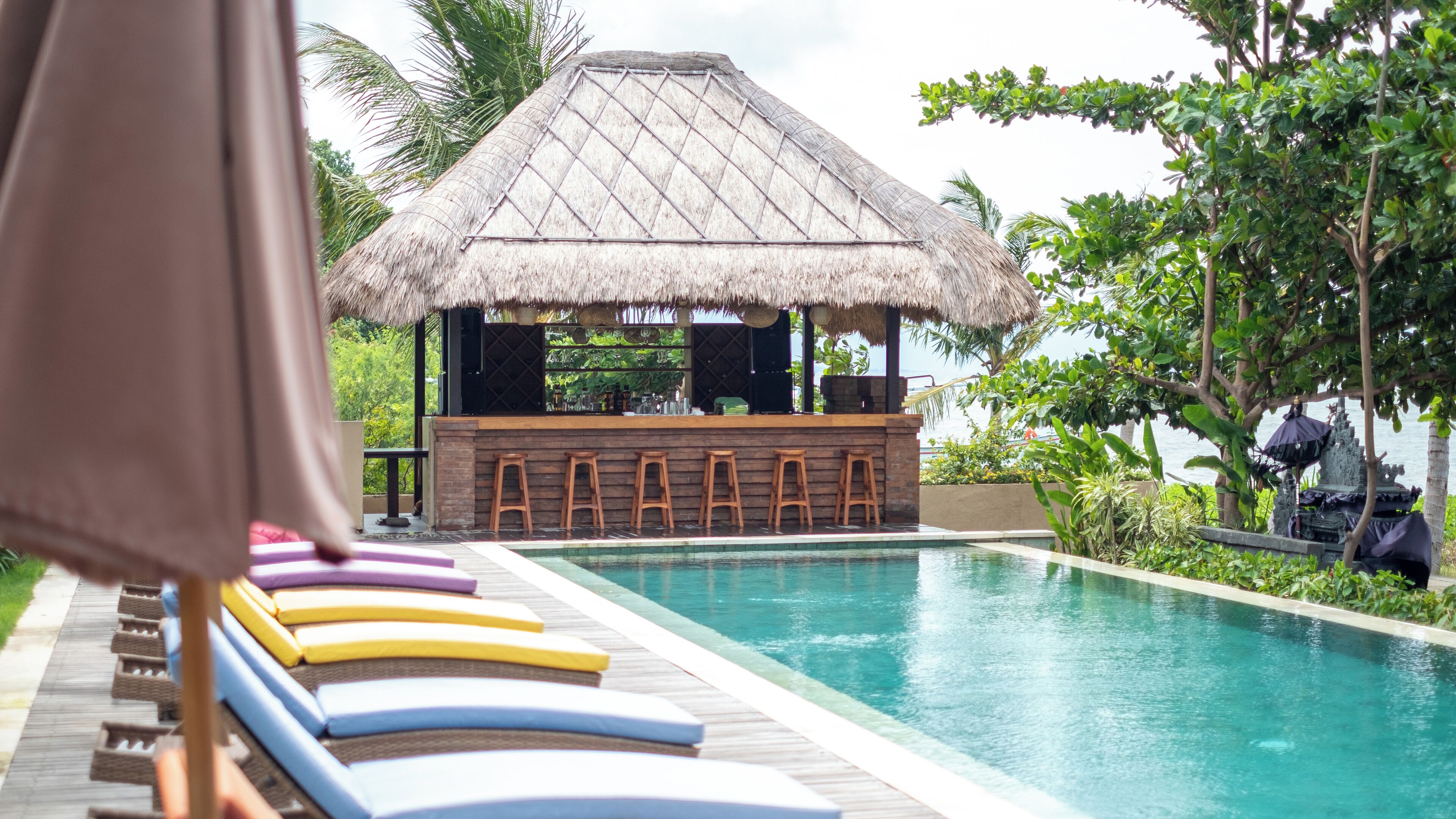 Un área junto a la piscina con sillones de descanso, un bar y una estructura con techo de paja.