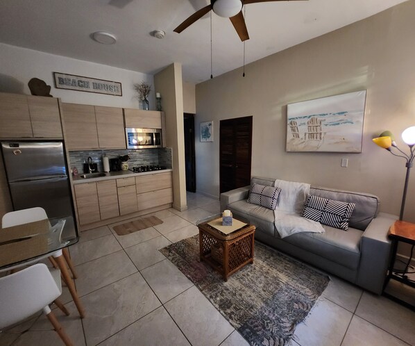 A modern living room with a grey sofa, a coffee table, and a kitchen area with wooden cabinets and a refrigerator.