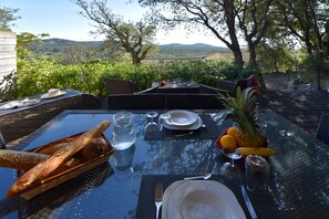 An outdoor dining table set with plates, cutlery, glasses, and a bottle, surrounded by greenery and a view of hills. - Villa-Superior-Private Bathroom-Sea View-Villa 4 - 6 personnes (Sainte-Lucie de Porto-Vecchio)