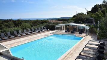 Une piscine dotée de chaises longues et d’un espace clos par des parois de verre, entourée de verdure et offrant une vue imprenable sur un paysage pittoresque.