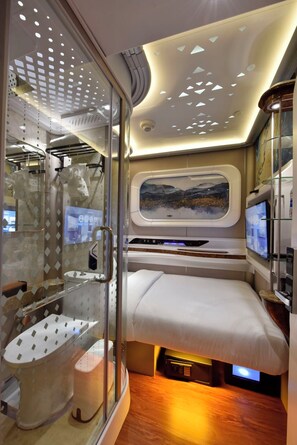 A compact, modern room with a bed, a small seating area, a window with a scenic view, and a glass partition with a shower area. - Bahrain Airport Hotel Airside Hotel for Transiting and Departing Passengers only (Muharraq)