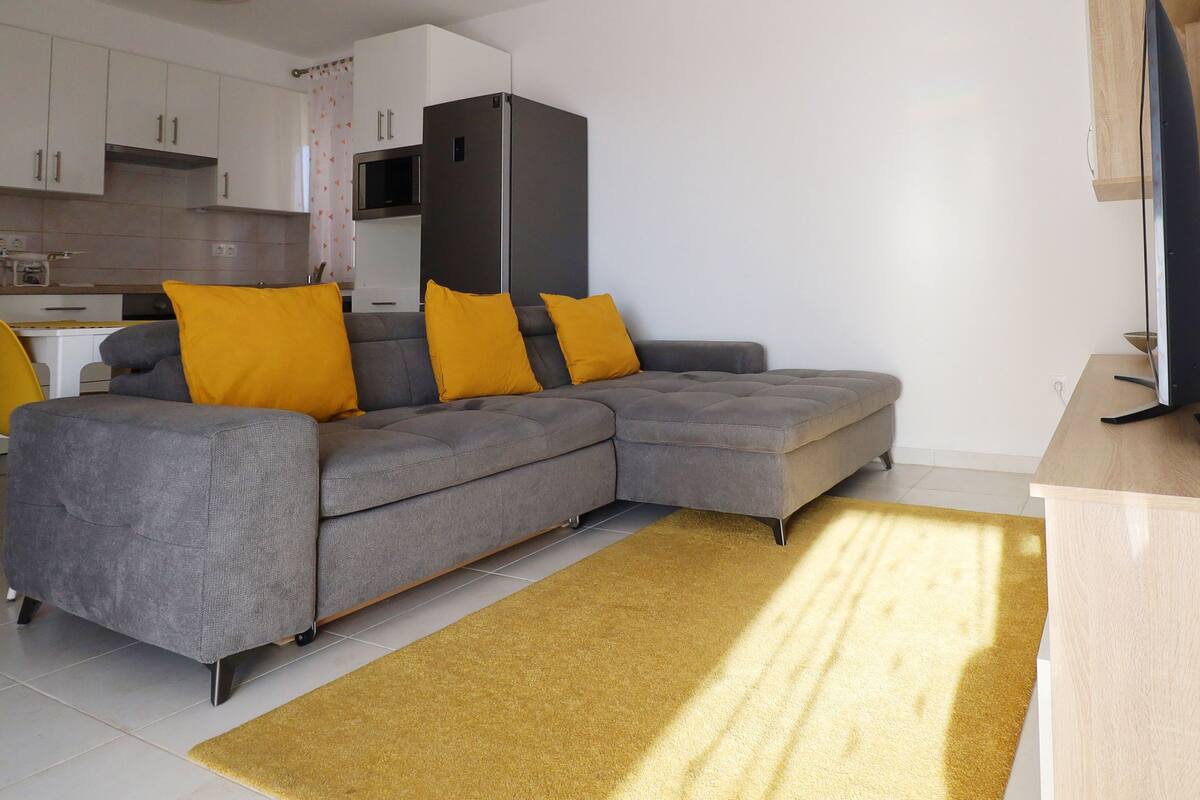 Ein modernes Wohnzimmer mit einer grauen Eckcouch, zwei gelben Kissen, einem Holztisch und einem gelben Teppich.