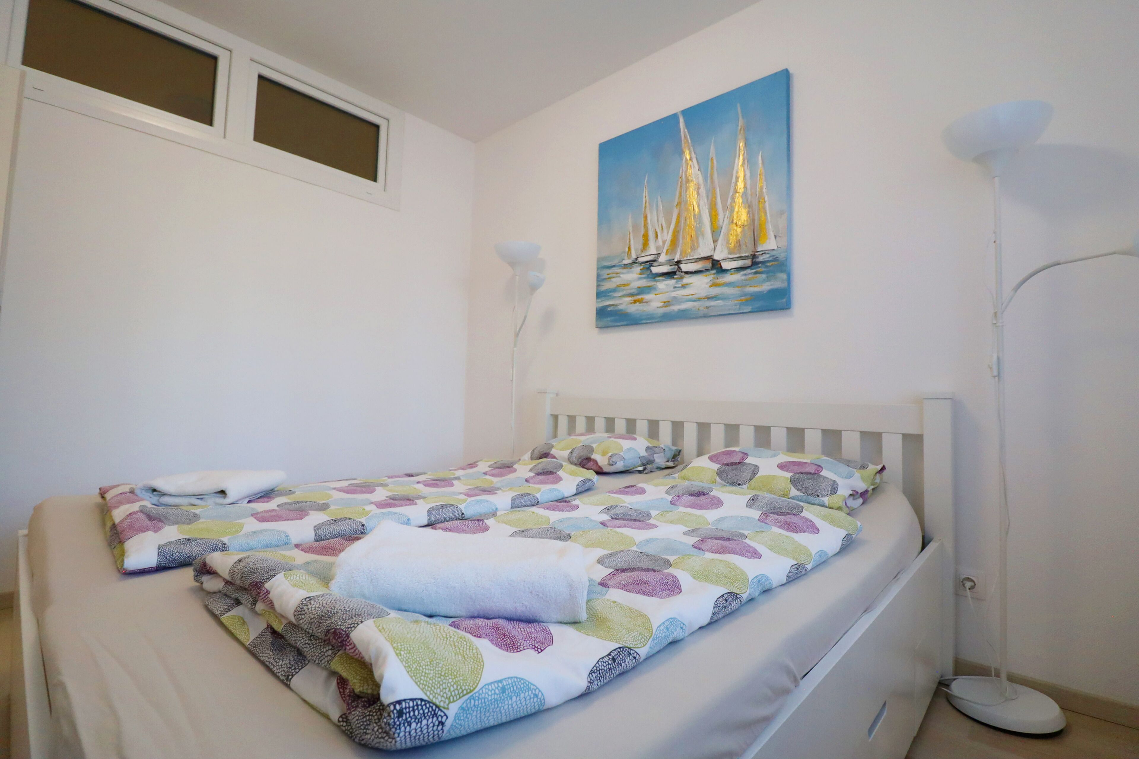 A neatly made bed with a colorful patterned bedspread, a painting of sailboats on the wall, and a floor lamp beside the bed.