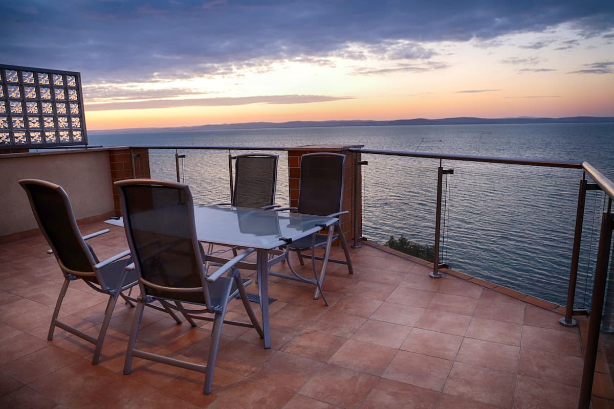 Eine Terrasse mit einem Glastisch und Stühlen, mit Blick auf ein Gewässer und das entfernte Umland.