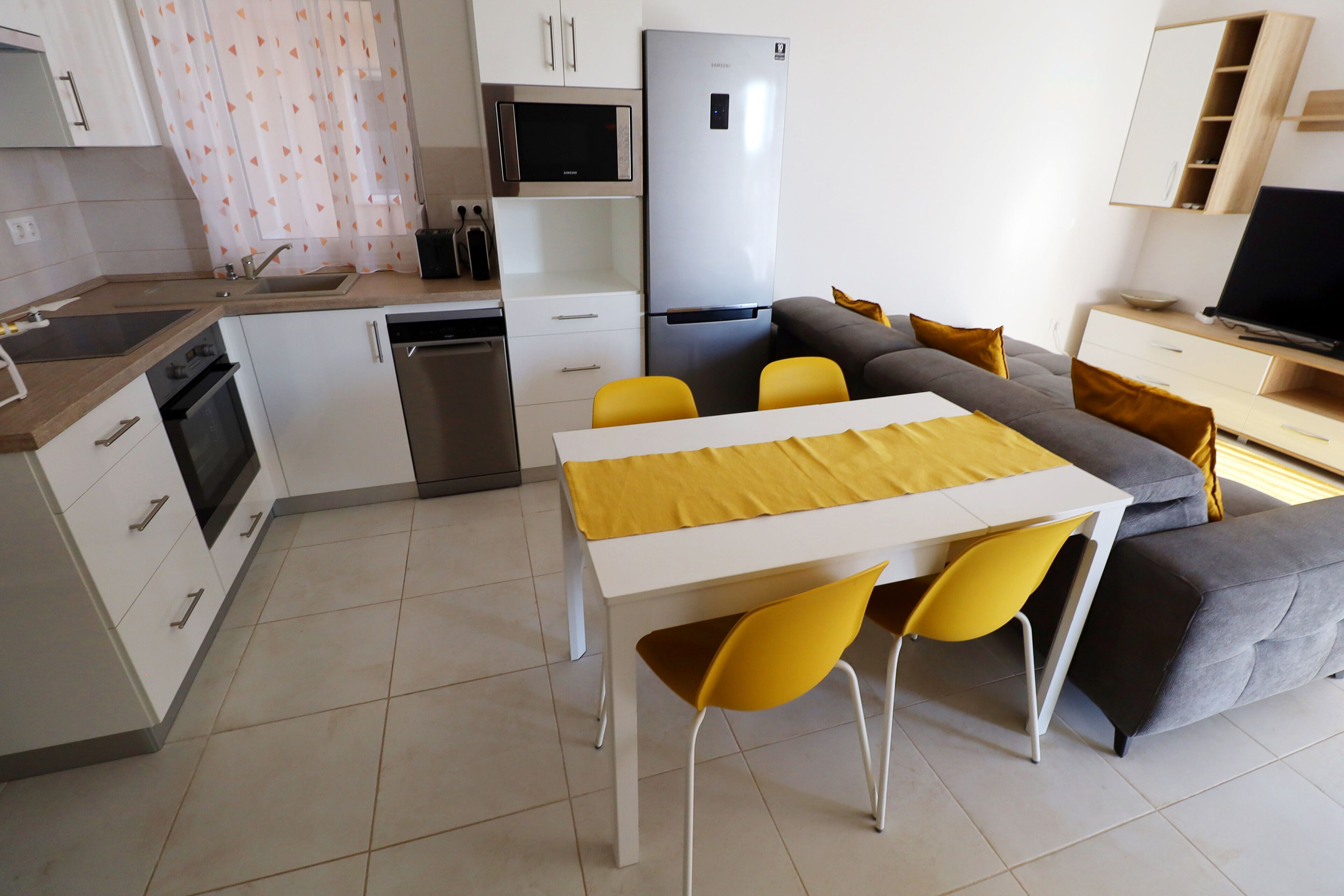 A modern kitchen with white cabinets, a stainless steel refrigerator, a microwave, and a sink. There is a dining area with a white table and yellow chairs.