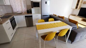 Una cocina moderna con armarios blancos, un refrigerador de acero inoxidable, un microondas y un lavamanos. Hay una zona de comedor con una mesa blanca y sillas amarillas.