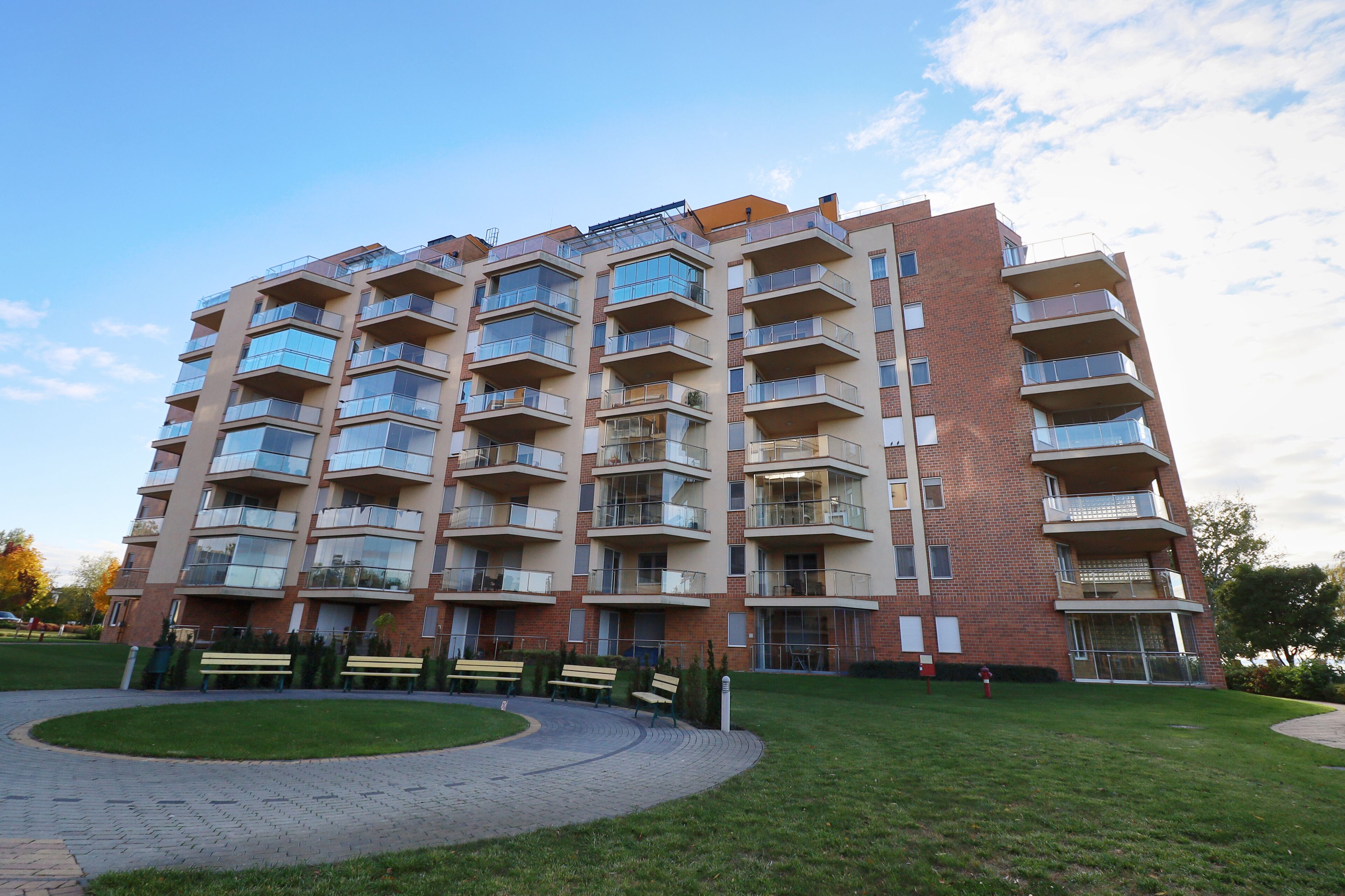A multi-story residential building with balconies, a paved courtyard, and benches.