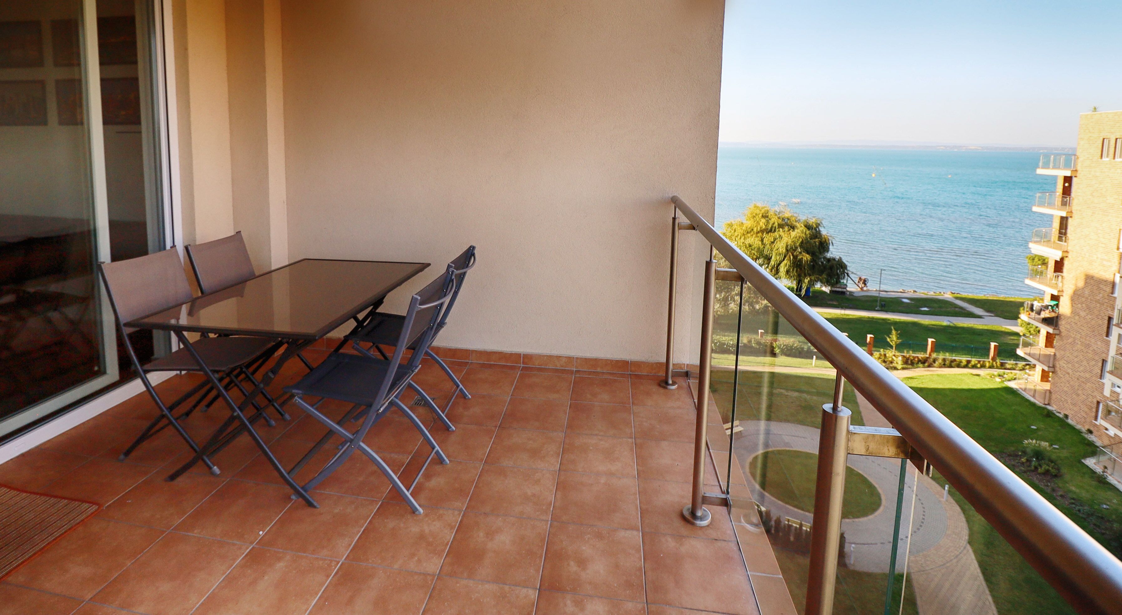 Un balcon avec une table et des chaises, une balustrade en verre et une vue sur la mer et les bâtiments.