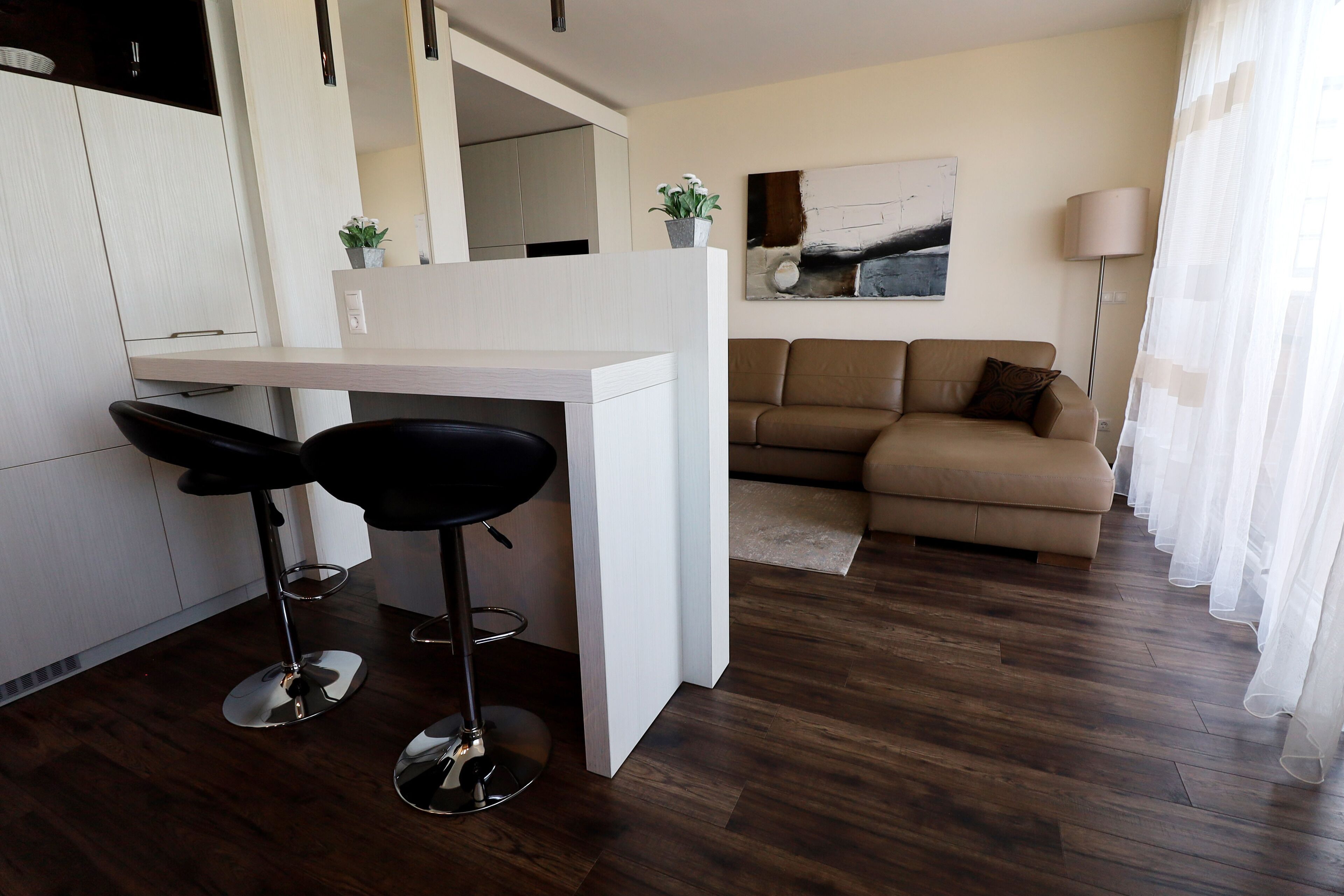 A modern kitchen with white cabinetry, two black bar stools, a white island, and a beige sofa in a living area with a wooden floor.