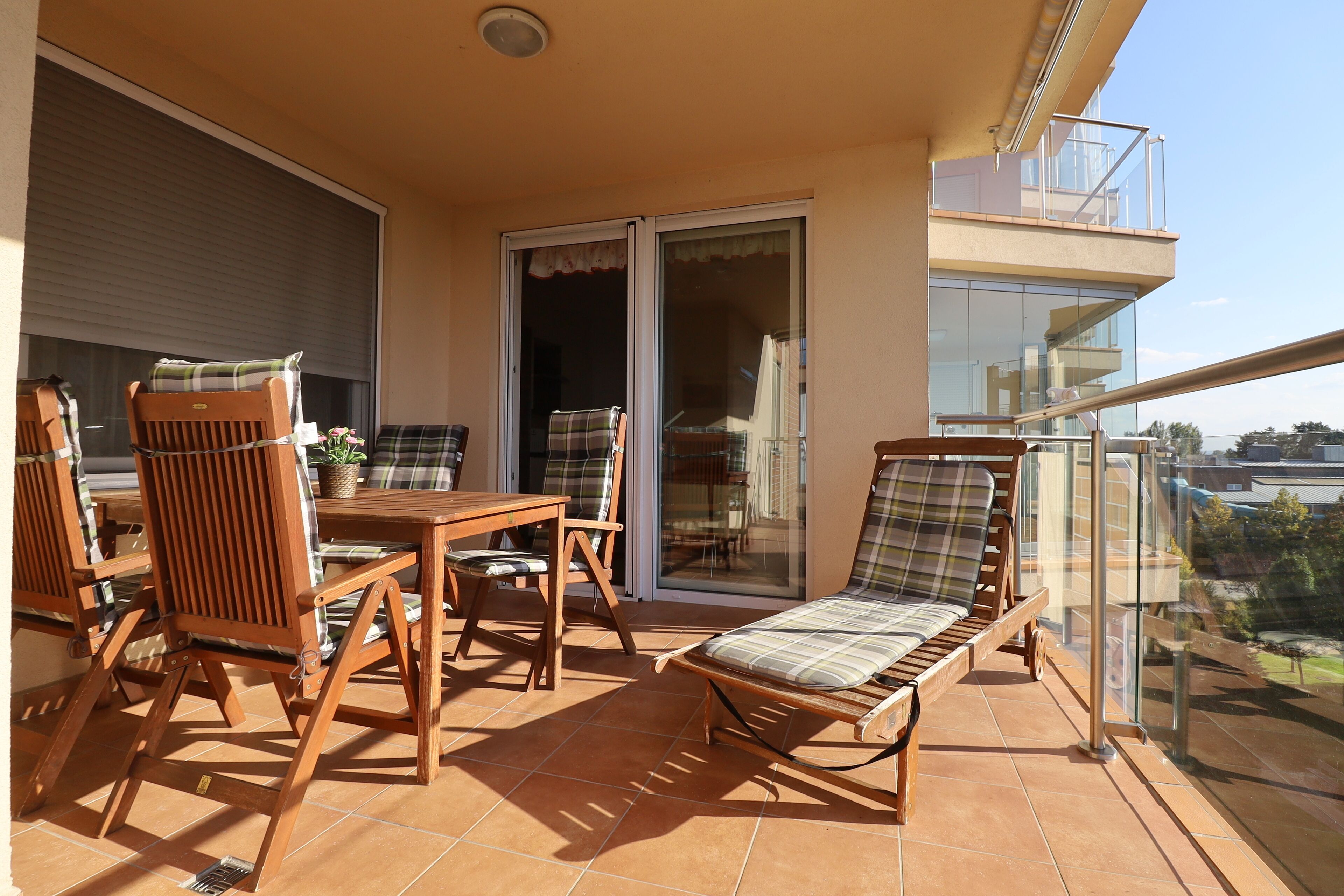 A balcony with a wooden table and chairs, a cushioned lounge chair, and a sliding glass door leading to an indoor area.