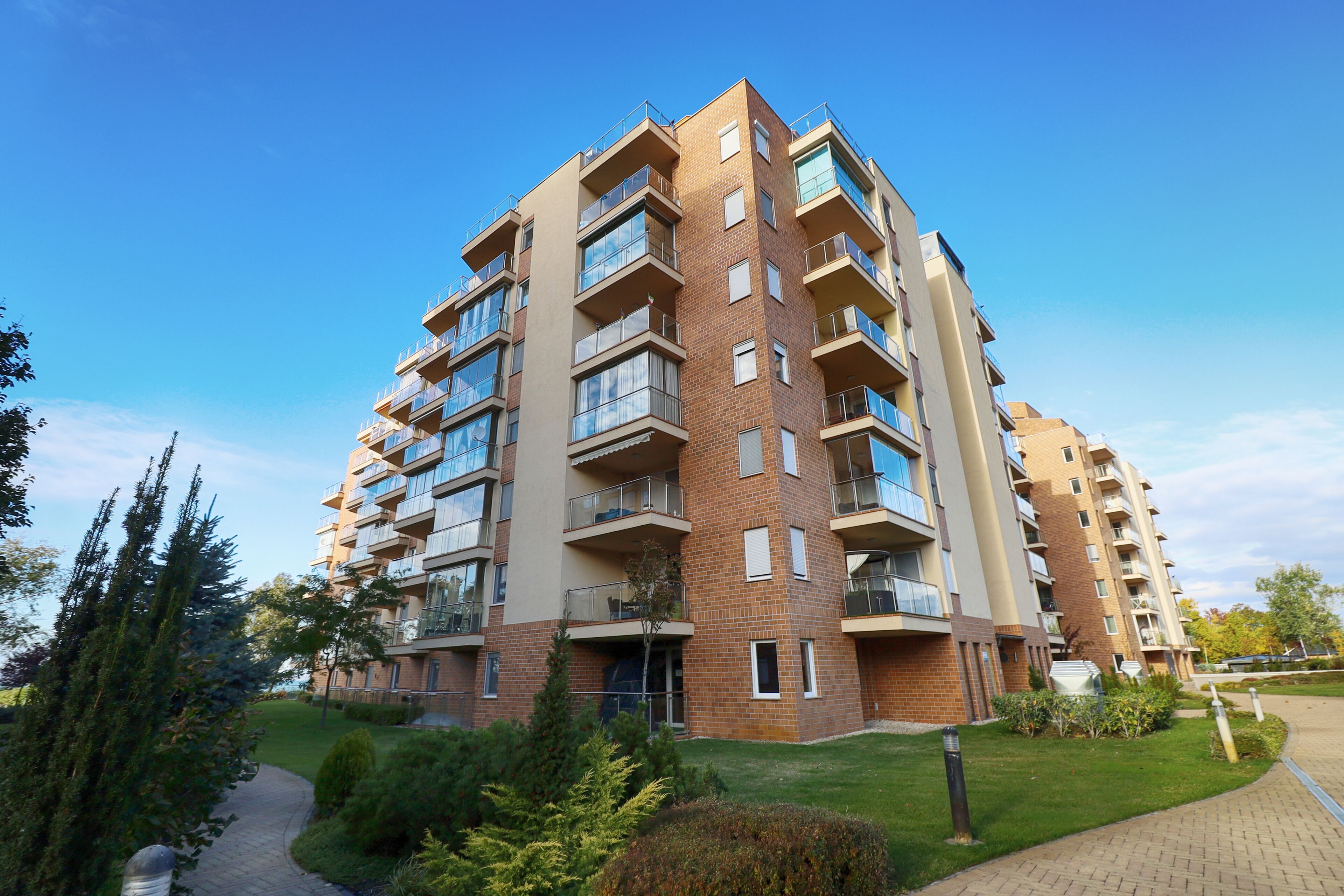 Edifício residencial de vários andares, com varandas, fachada de tijolos e um gramado bem cuidado.
