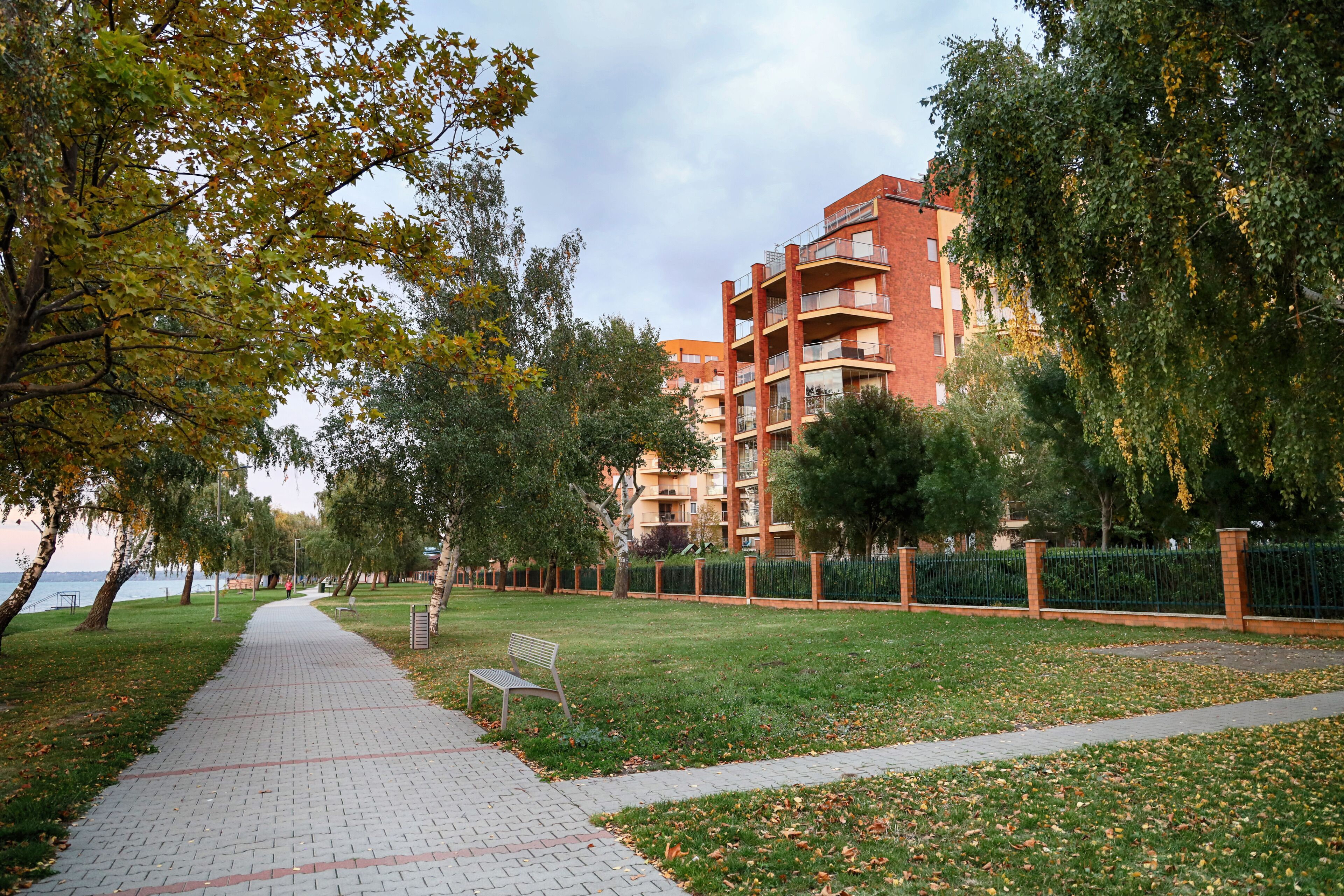 A park pathway with a bench, trees, and a building in the background.