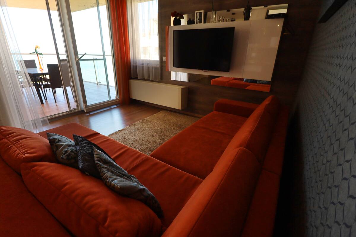 Ein modernes Wohnzimmer mit einem großen, orangefarbenen Sofa, einem flachbildfernseher an der Wand und einer Glastür, die auf einen Balkon führt.