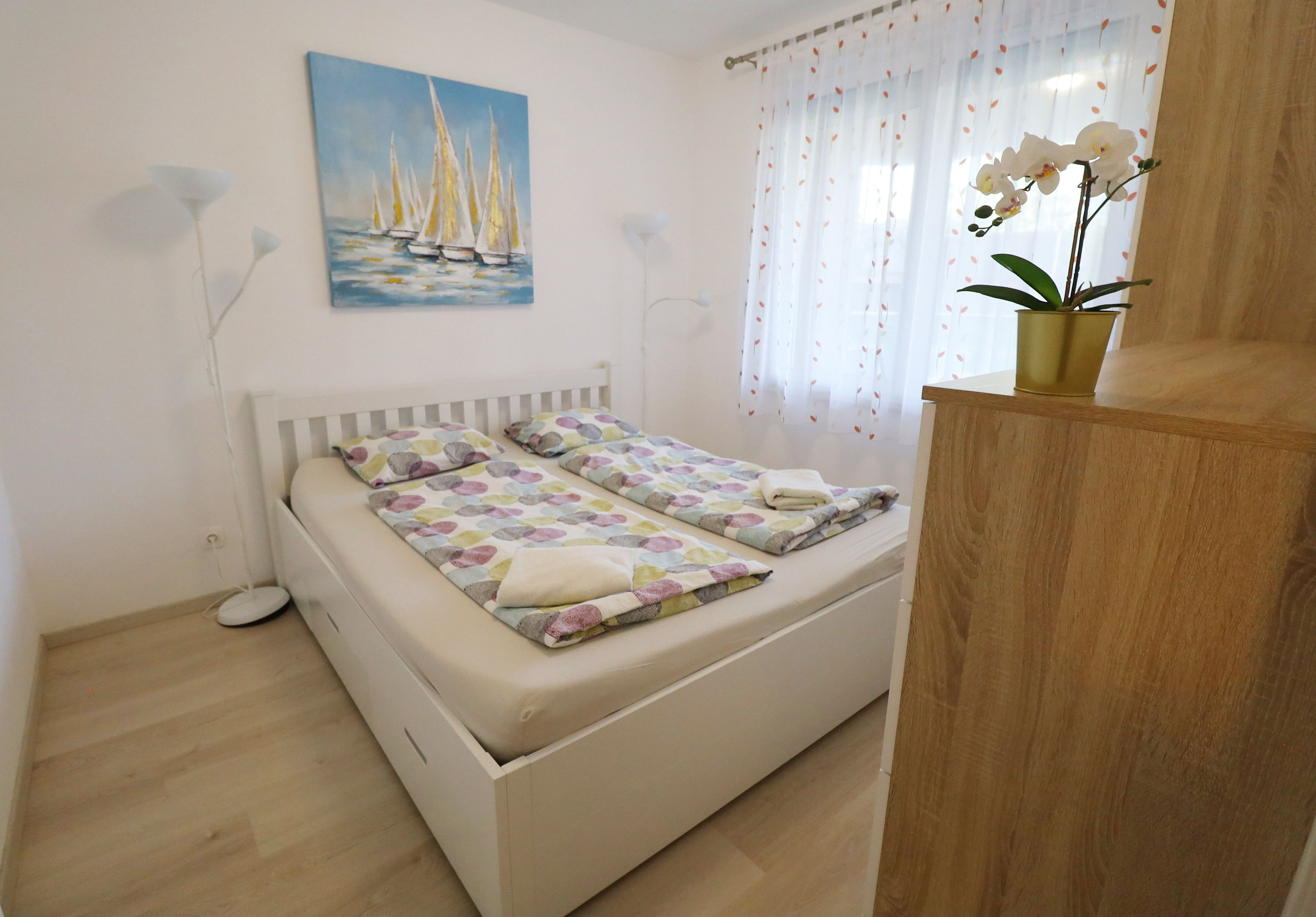 A bedroom with a bed, a painting of sailboats, a potted orchid, and a wooden cabinet.