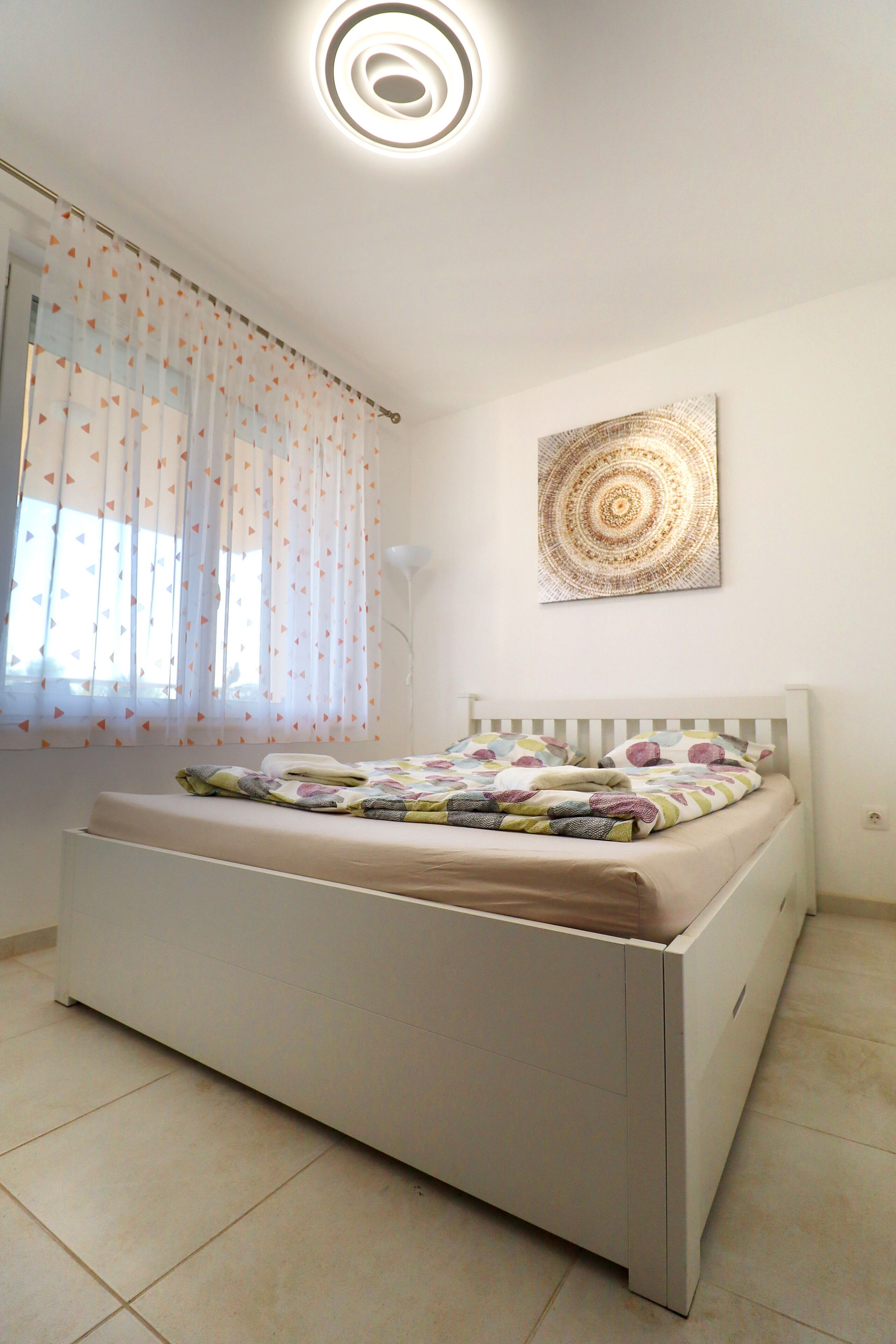 Un dormitorio con una cama, una ventana con cortinas translúcidas, una lámpara fijada a la pared y una obra de arte circular en la pared.