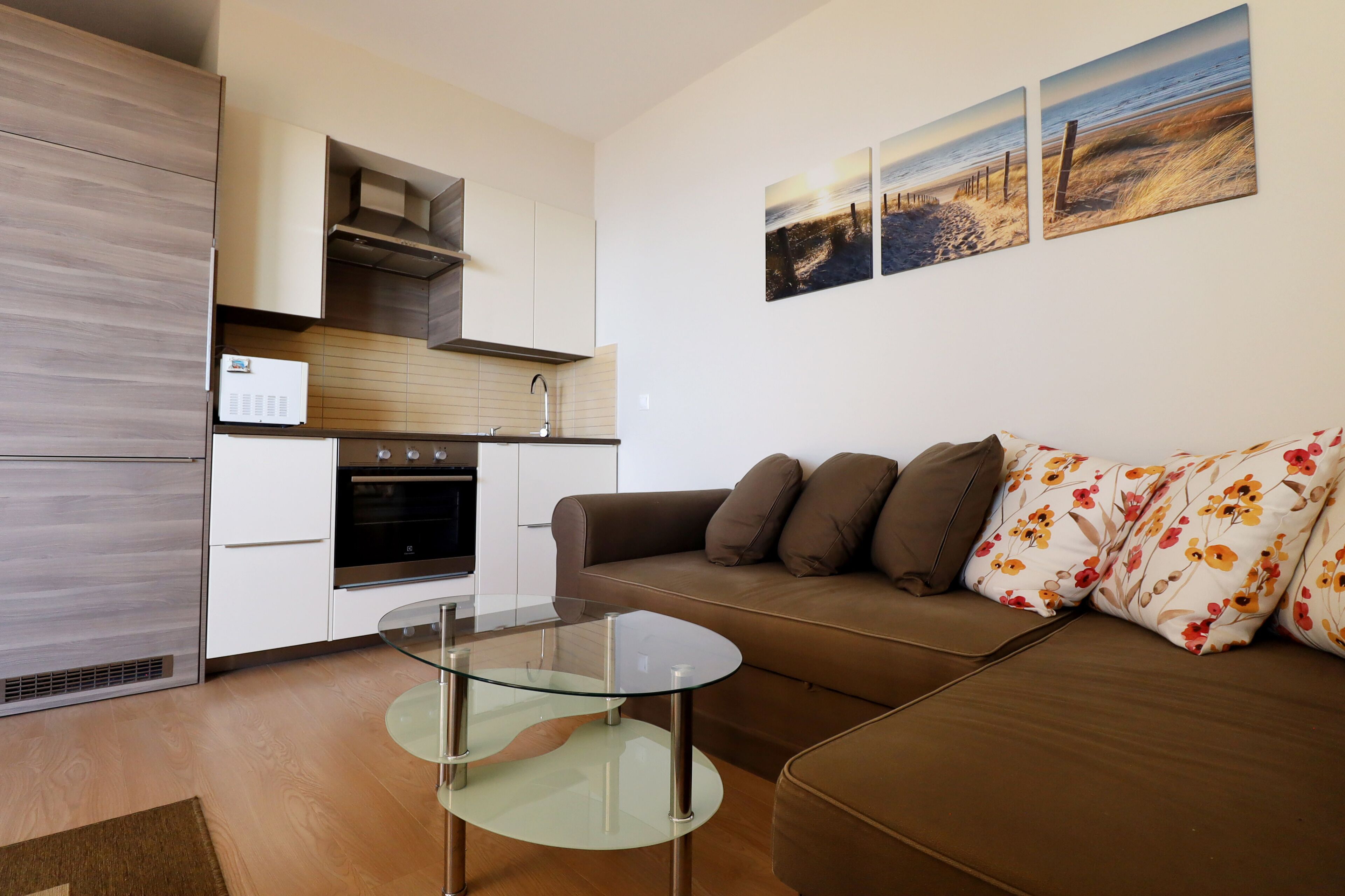 A modern kitchen with white cabinets, a stainless steel range hood, and a built-in oven. There is a glass coffee table in the living area, a brown sofa with floral cushions, and a wall-mounted artwork depicting a beach scene.