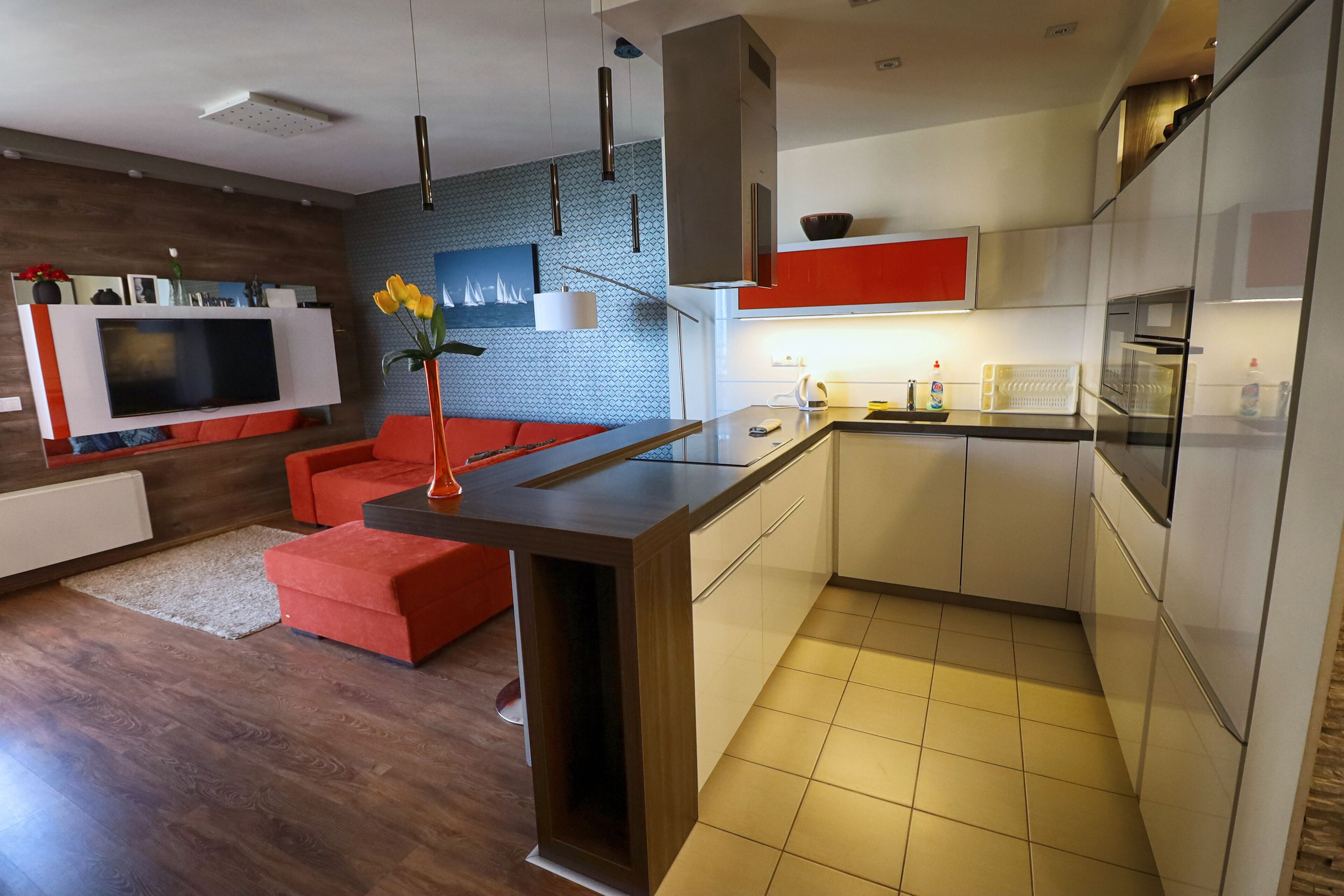 A modern kitchen with a central island, a dining area with red furniture, and a living room with a TV and red cushions.
