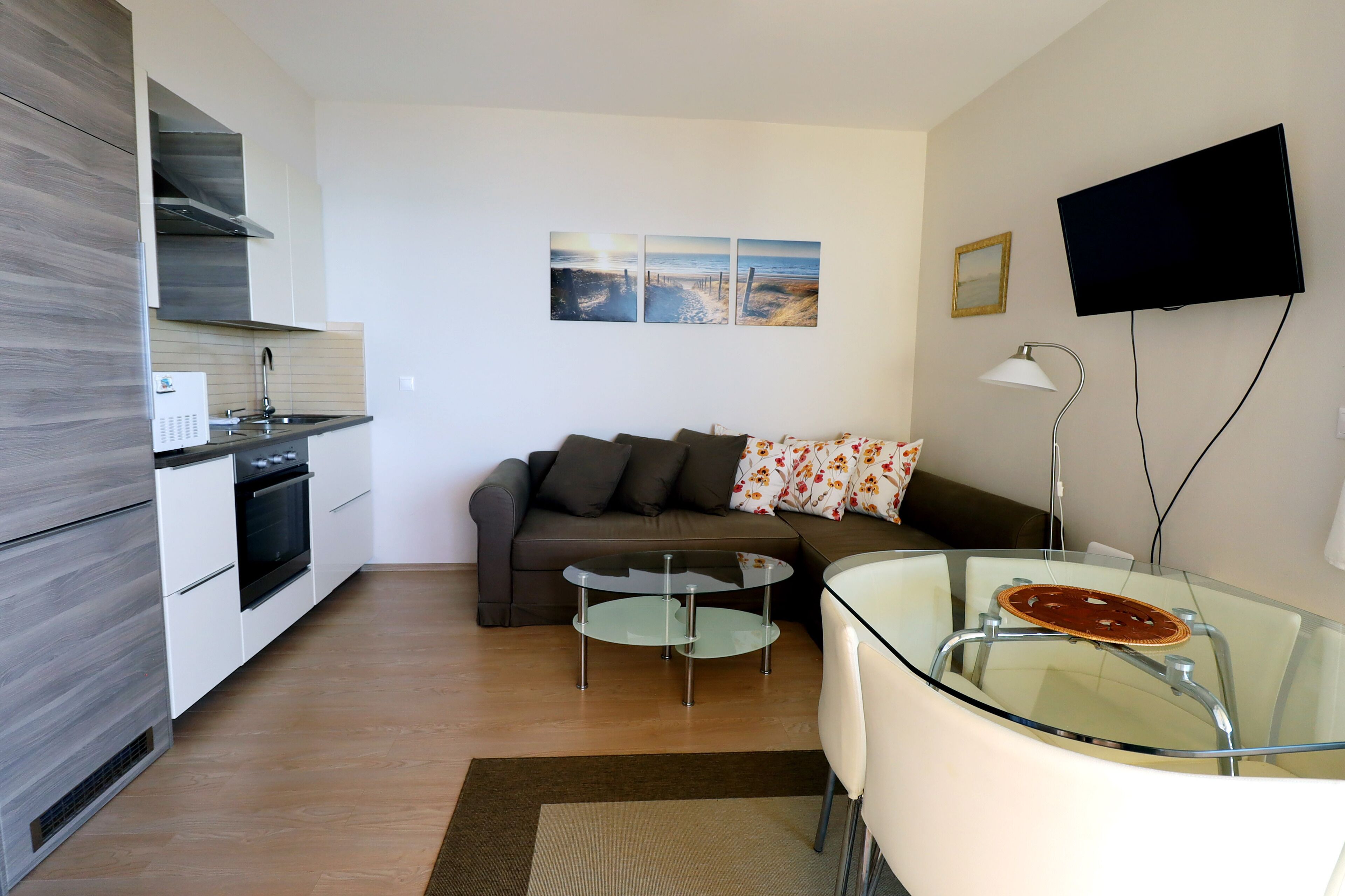 A modern kitchen with built-in appliances, a dining area with a glass table, a sofa with floral cushions, and a flat-screen TV mounted on the wall.