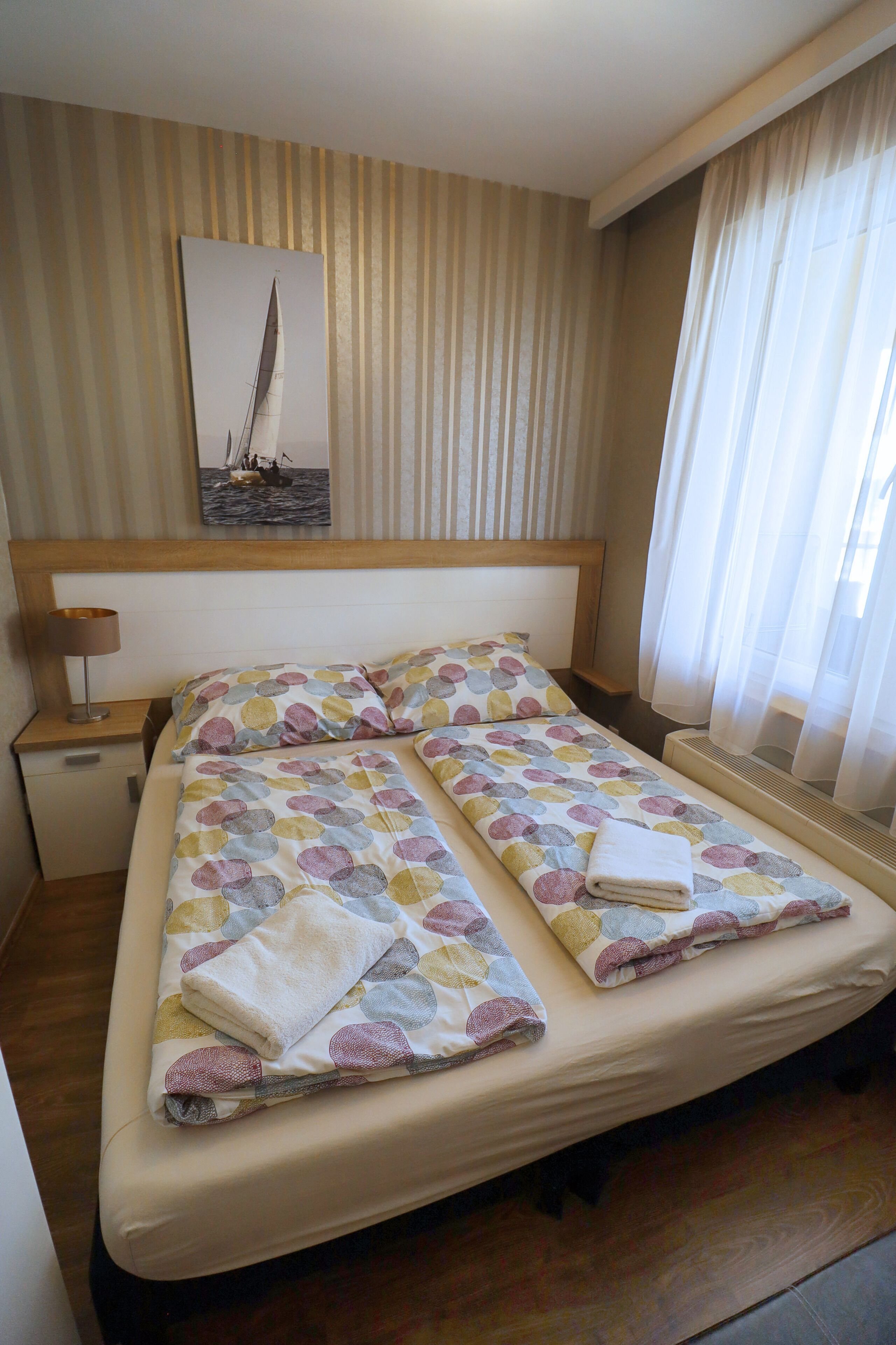 A hotel room with a bed, a nightstand, a lamp, a painting of a sailboat, and a window with curtains.