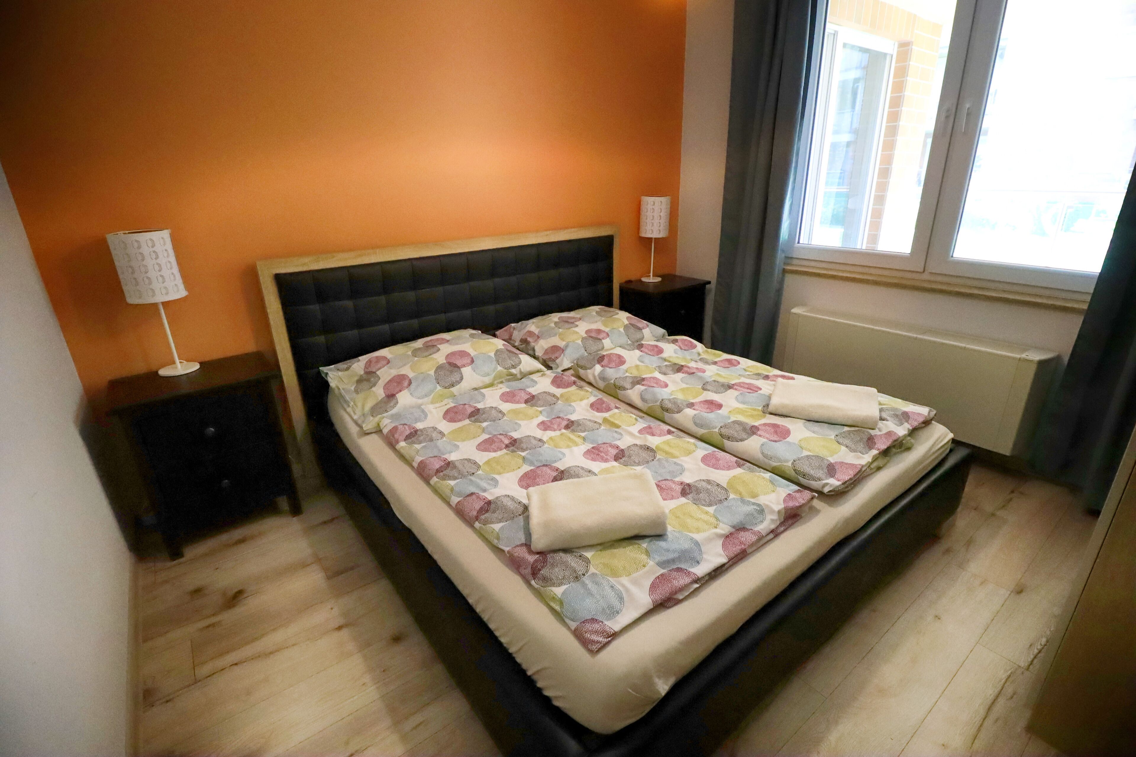 A double bed with a patterned bedspread, two bedside tables with lamps, and a window with curtains.