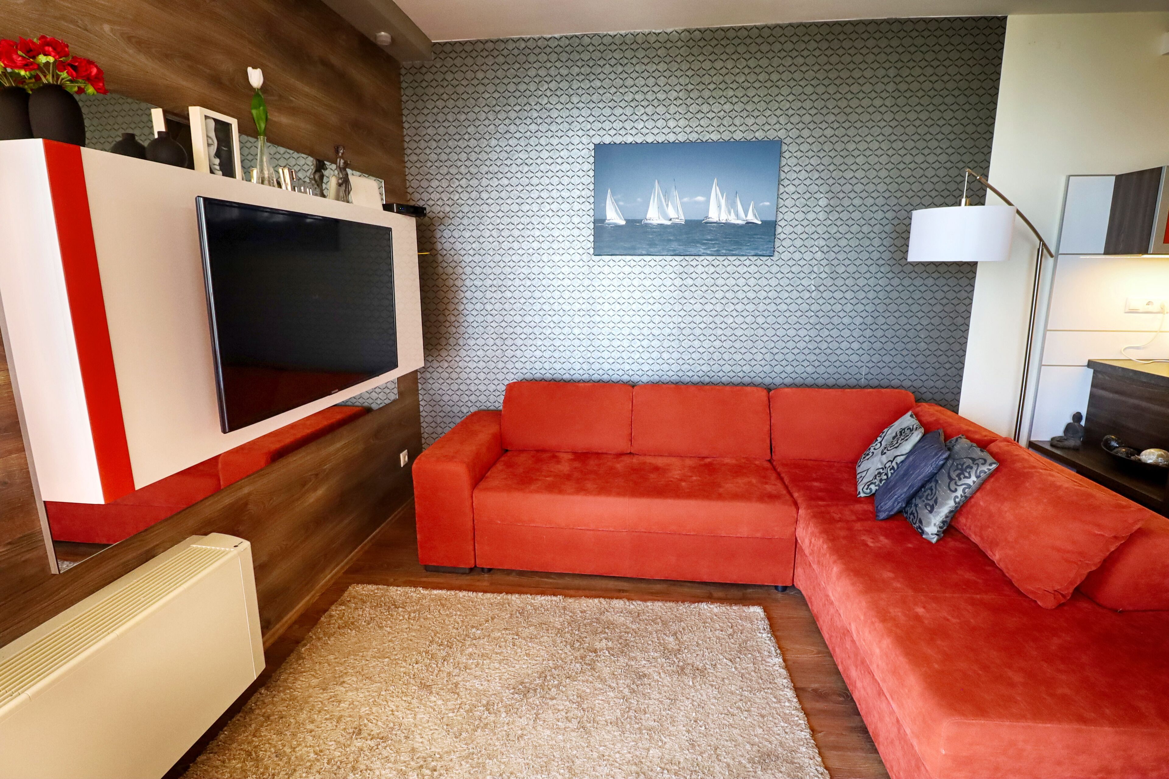 Una sala de estar moderna con un sofá rojo, un televisor de pantalla plana, un papel tapiz estampado y un cuadro con barcos de vela.