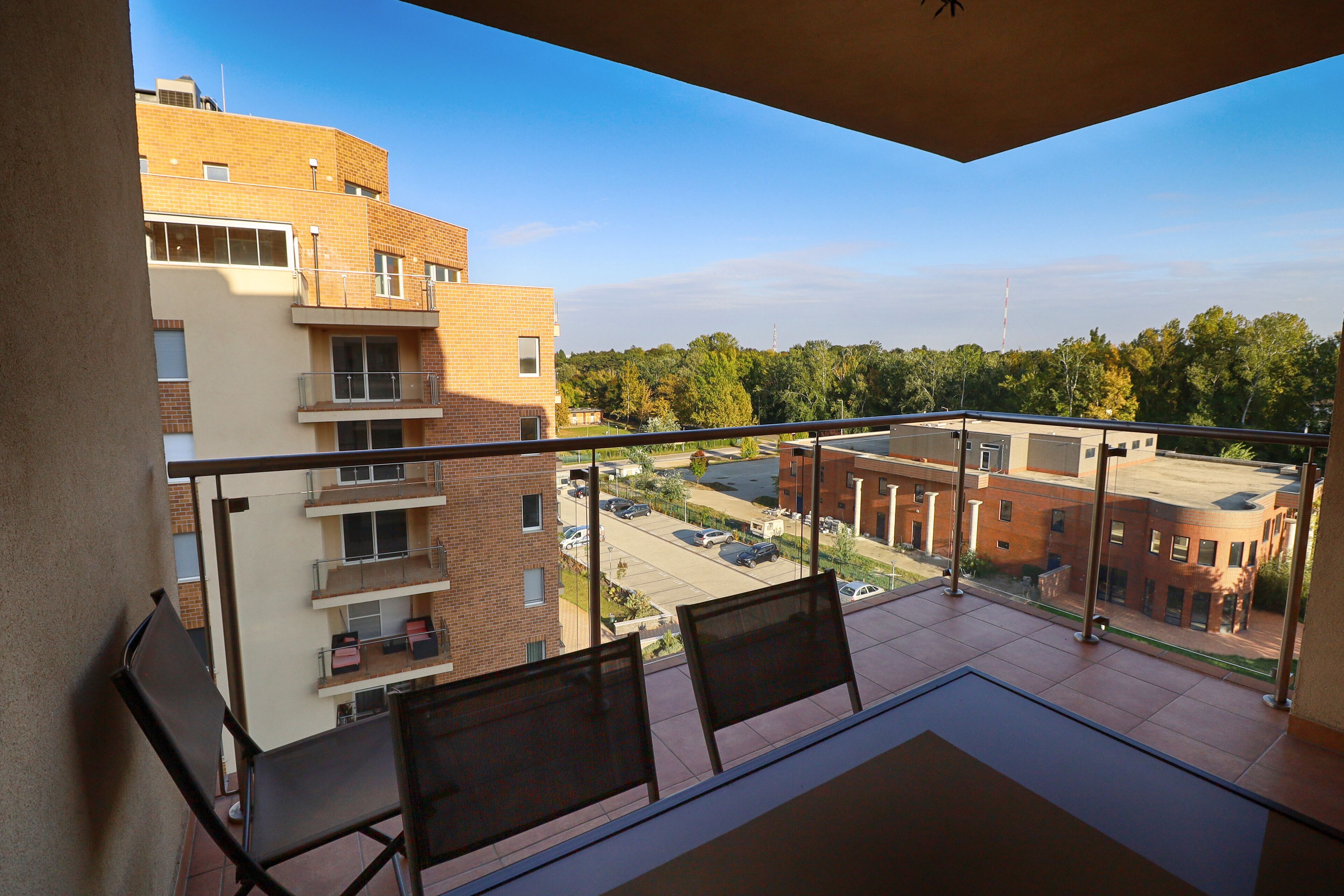 A balcony with two chairs, a table, and a view of a parking lot and buildings.